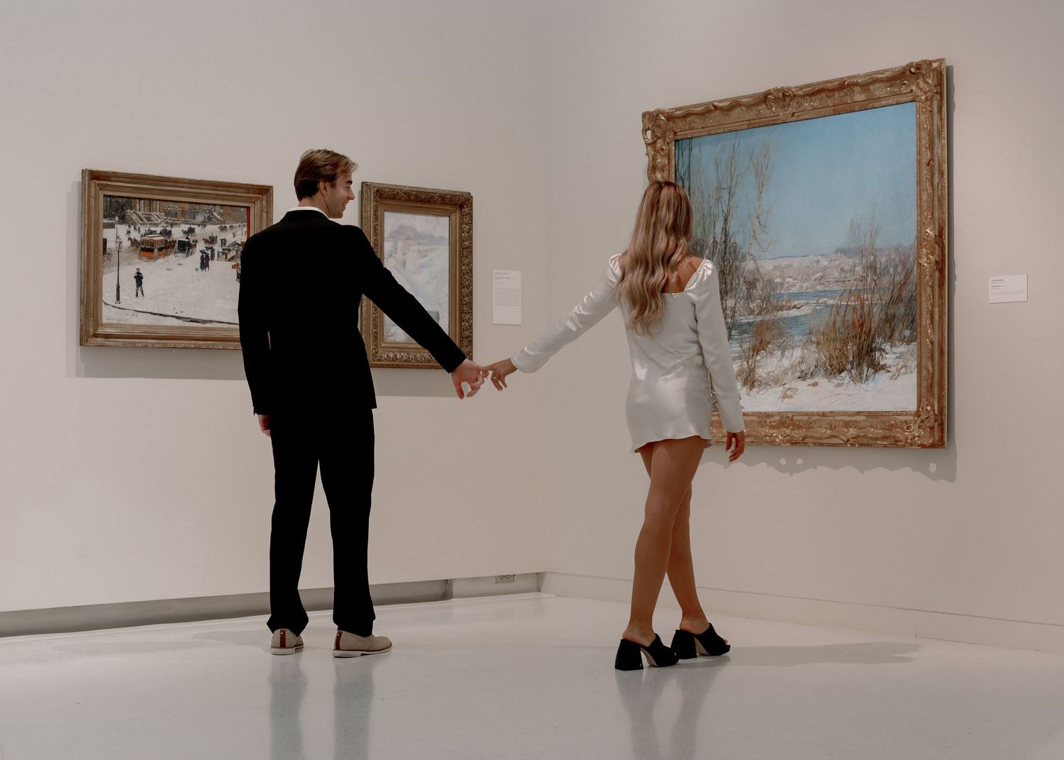 Two people holding hands while viewing artwork in a modern gallery with framed paintings mounted on white walls.