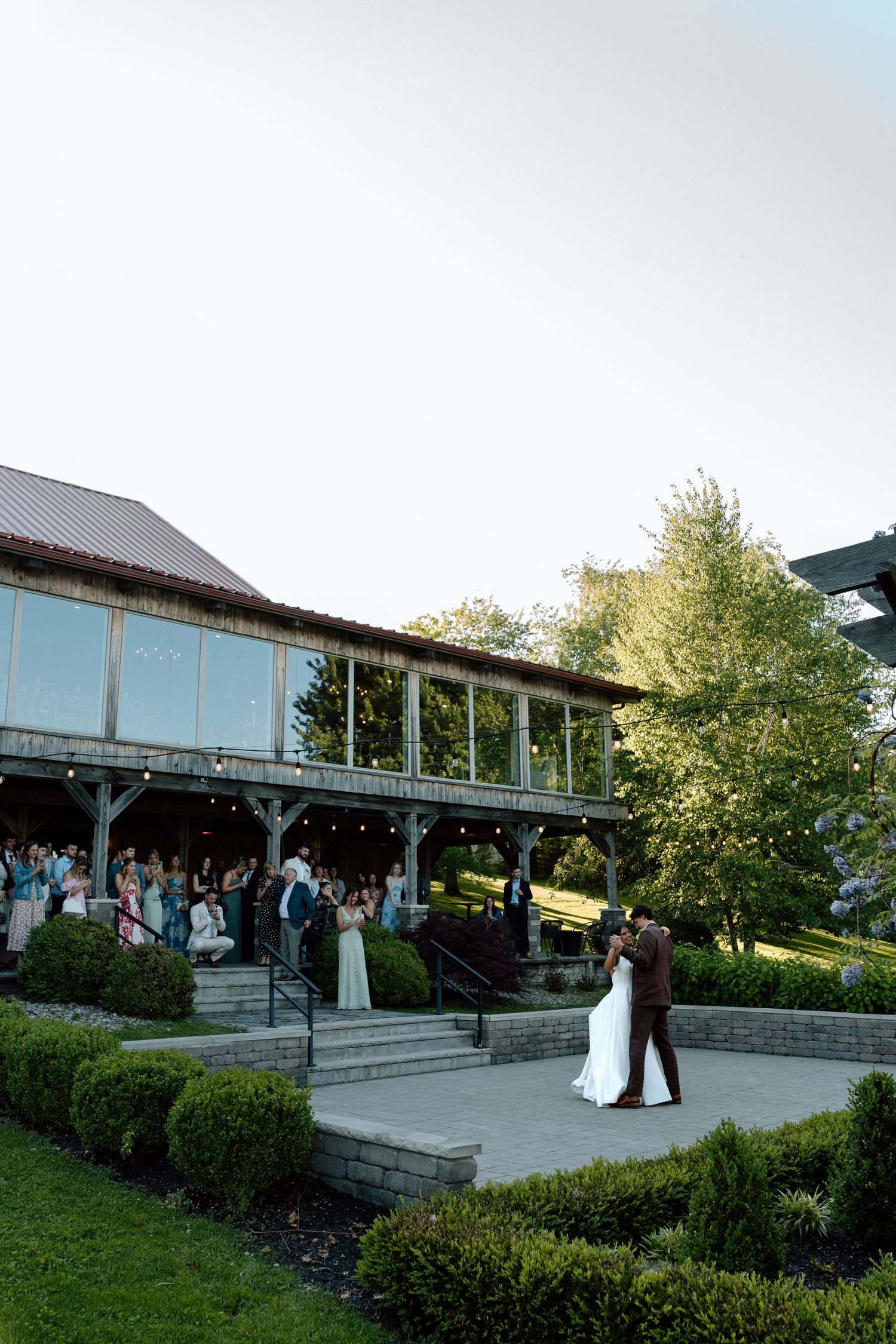 Bride and groom share first dance on outdoor patio at rustic wedding venue with guests watching from covered pavilion.
