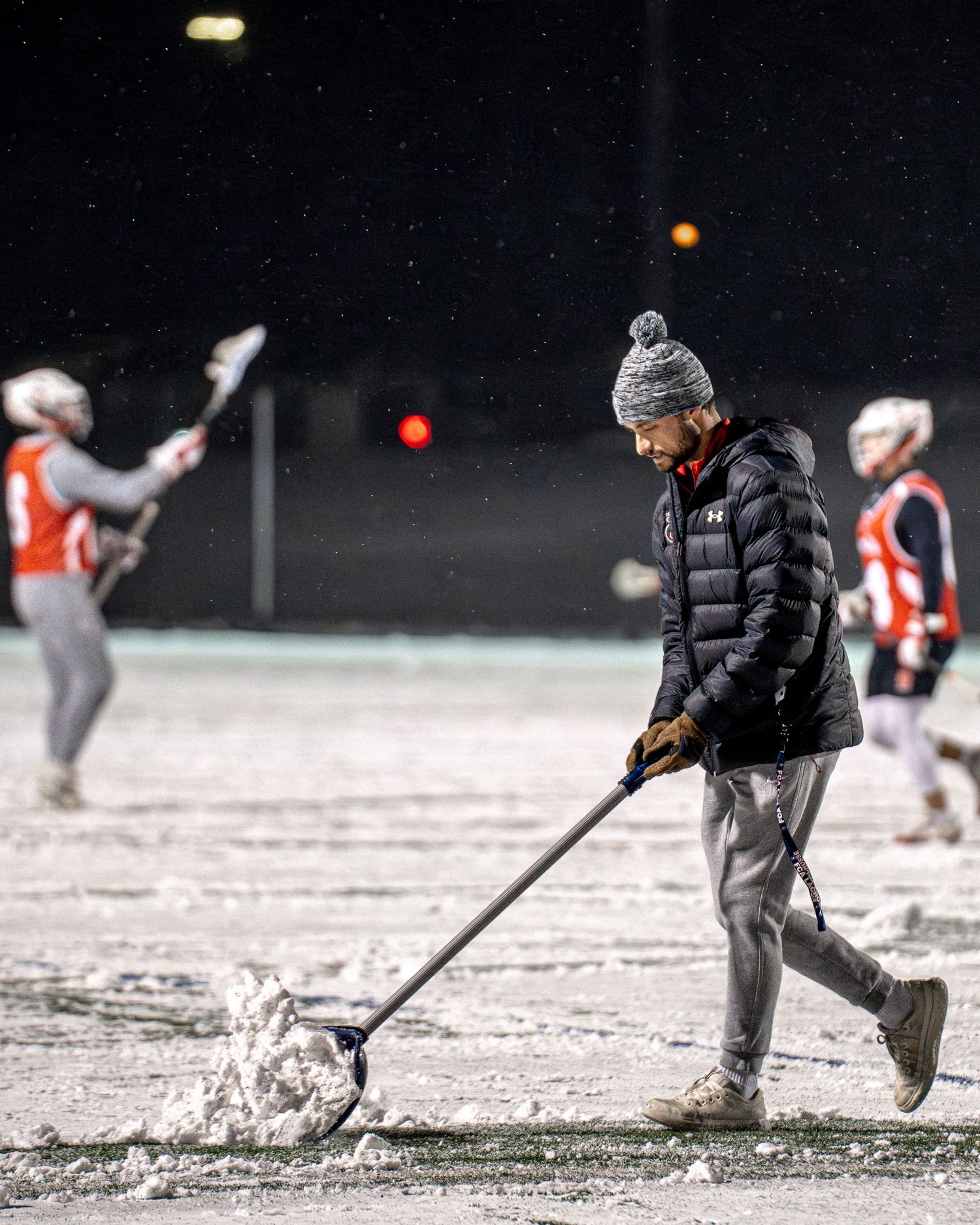Hockey players clear snow from an outdoor ice rink during a winter game in snowy conditions.