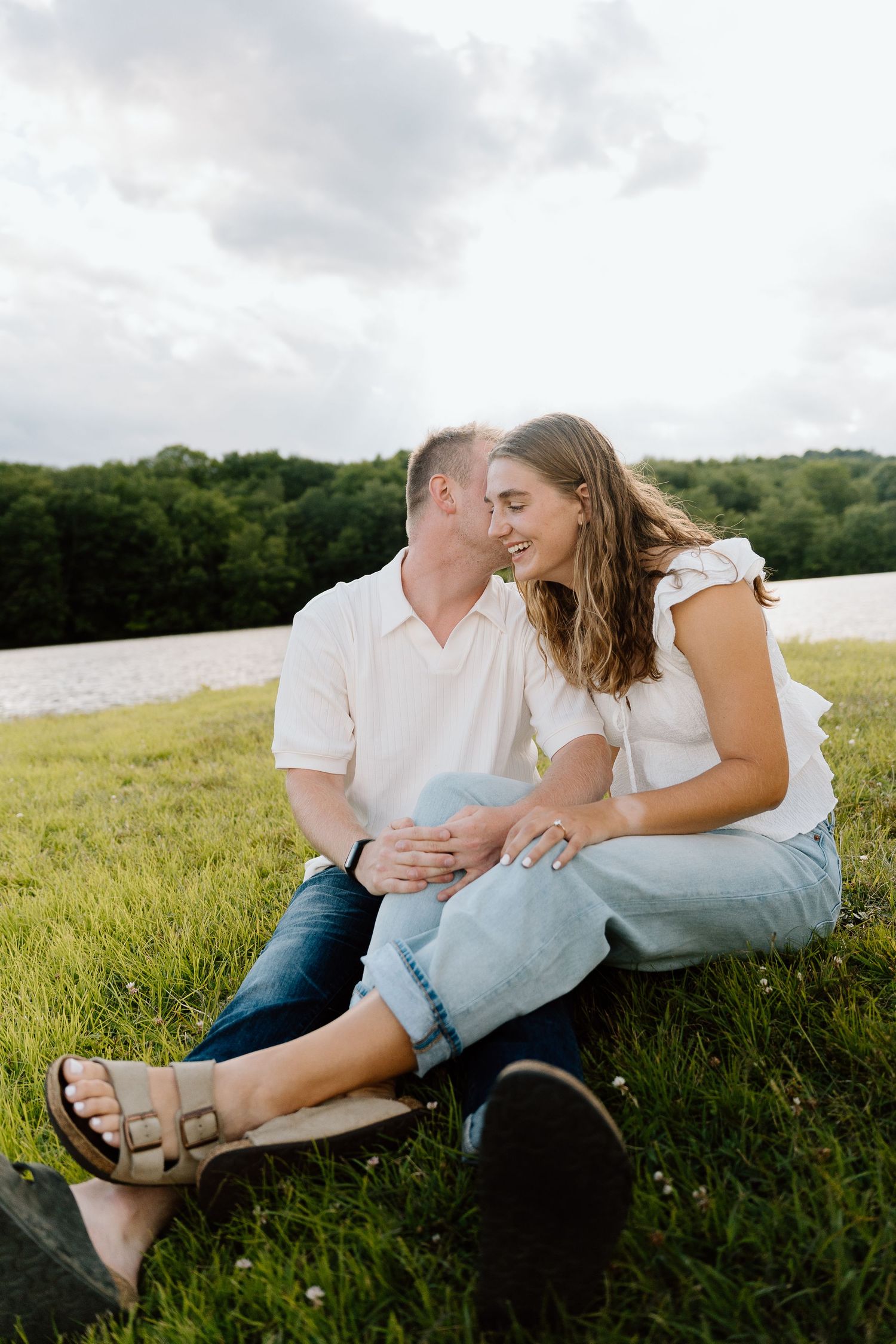 Couple sitting on green grass near a lake, sharing a romantic moment, dressed in casual white tops and jeans.