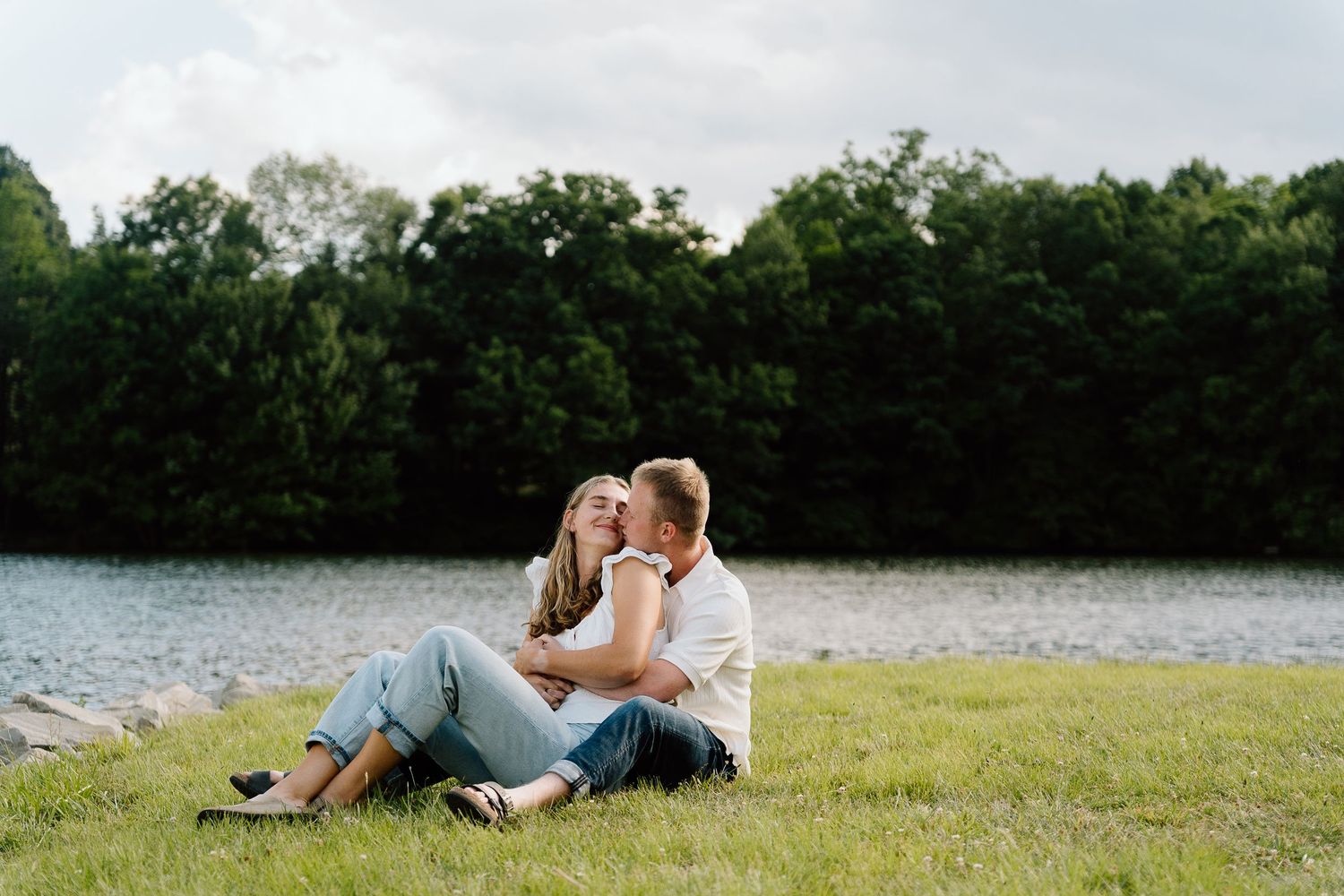 Couple sitting on grass by a lake, embracing romantically with lush green trees in the background.