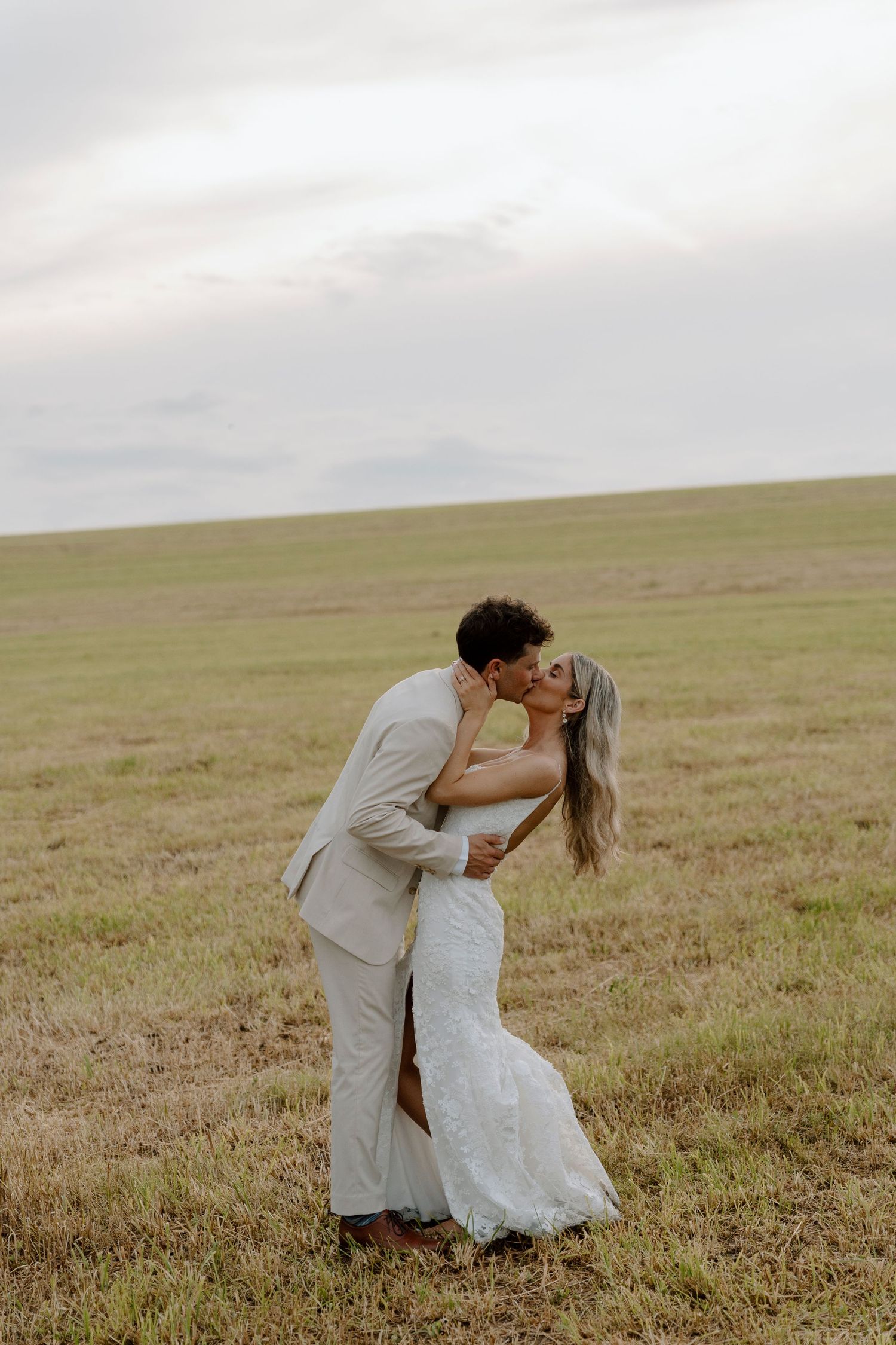 Couple in wedding attire share intimate moment in open field during sunset.
