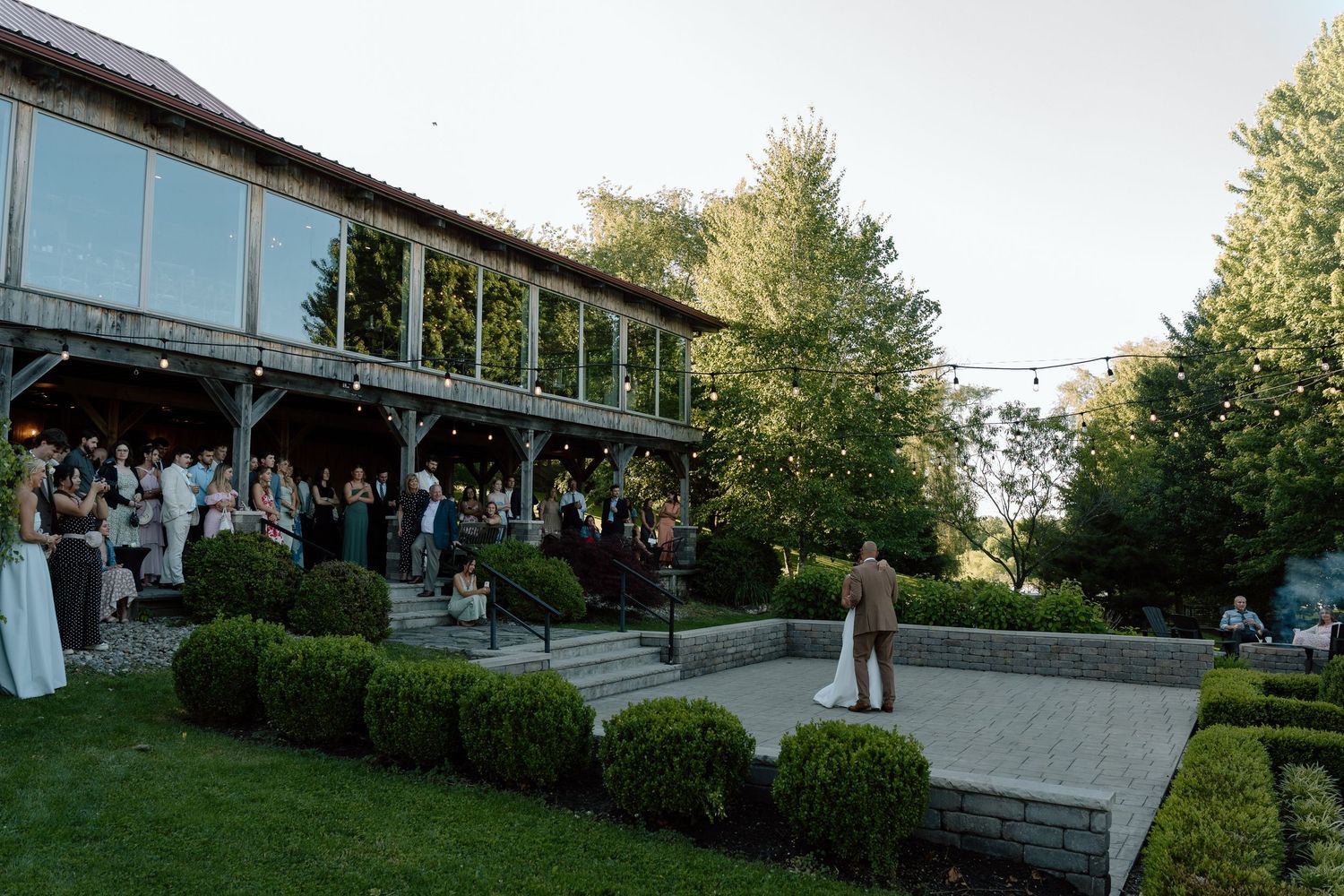 Elegant outdoor wedding reception on a patio with guests mingling near a rustic venue surrounded by lush greenery.