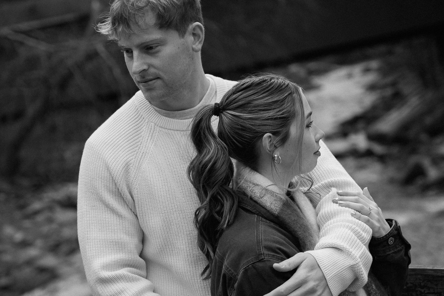 Black and white photo of a couple embracing outdoors, man in white sweater with arm around woman with ponytail.