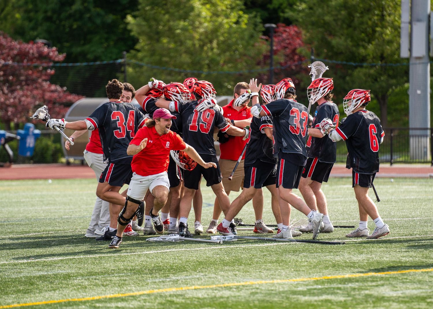 Grove City Men's Lacrosse celebrating in huddle formation on sports field wearing red and black uniforms.