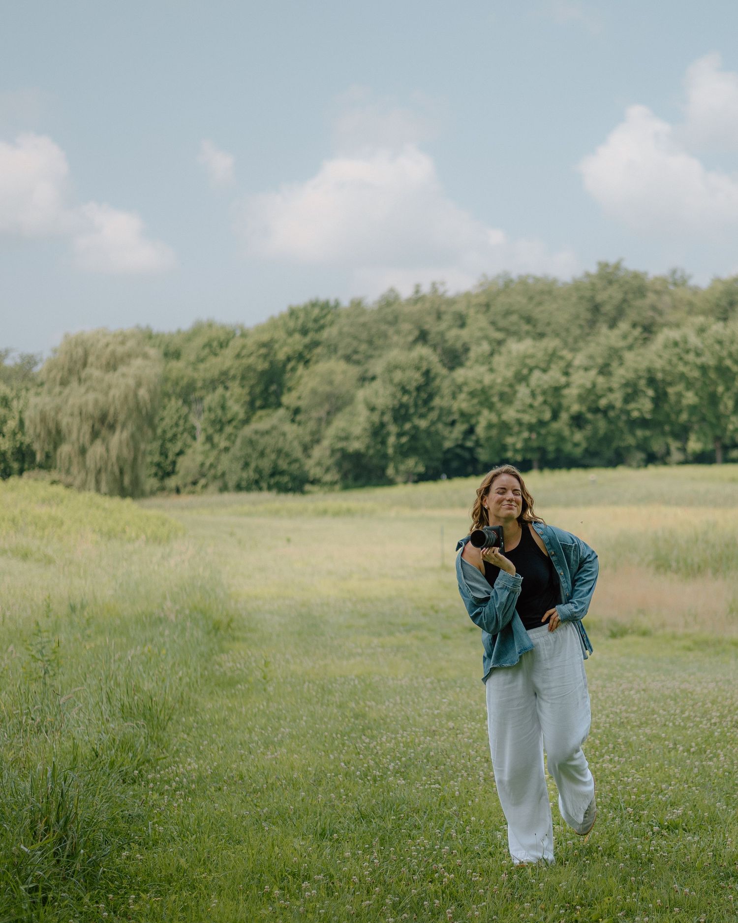 A person in white clothing dances and moves gracefully across a green meadow with trees in the background.