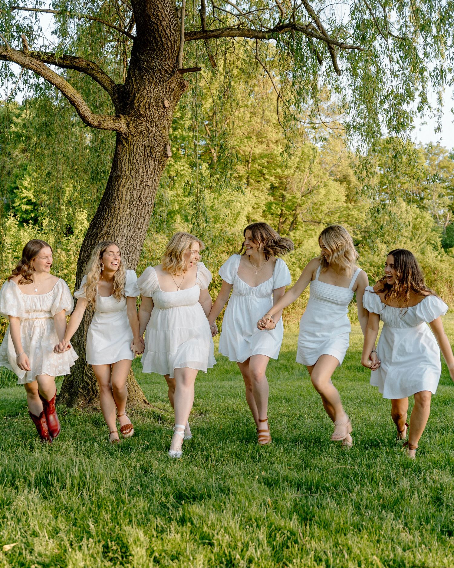 Group of friends sitting on grass under willow trees in white summer dresses.