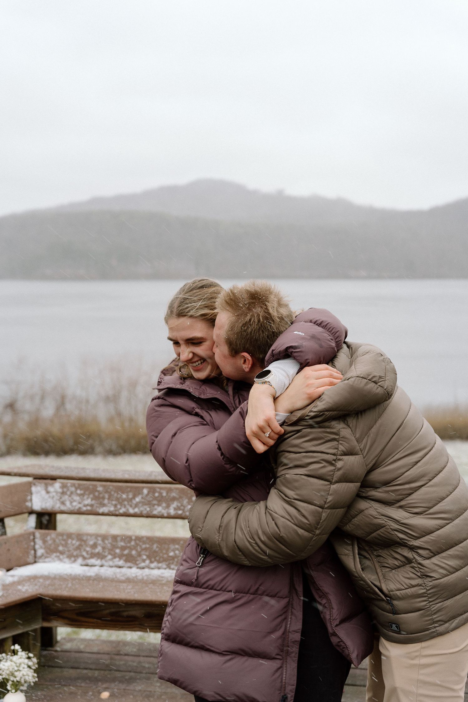 A couple embraces and laughs together on a wooden deck overlooking a misty lake and mountains.