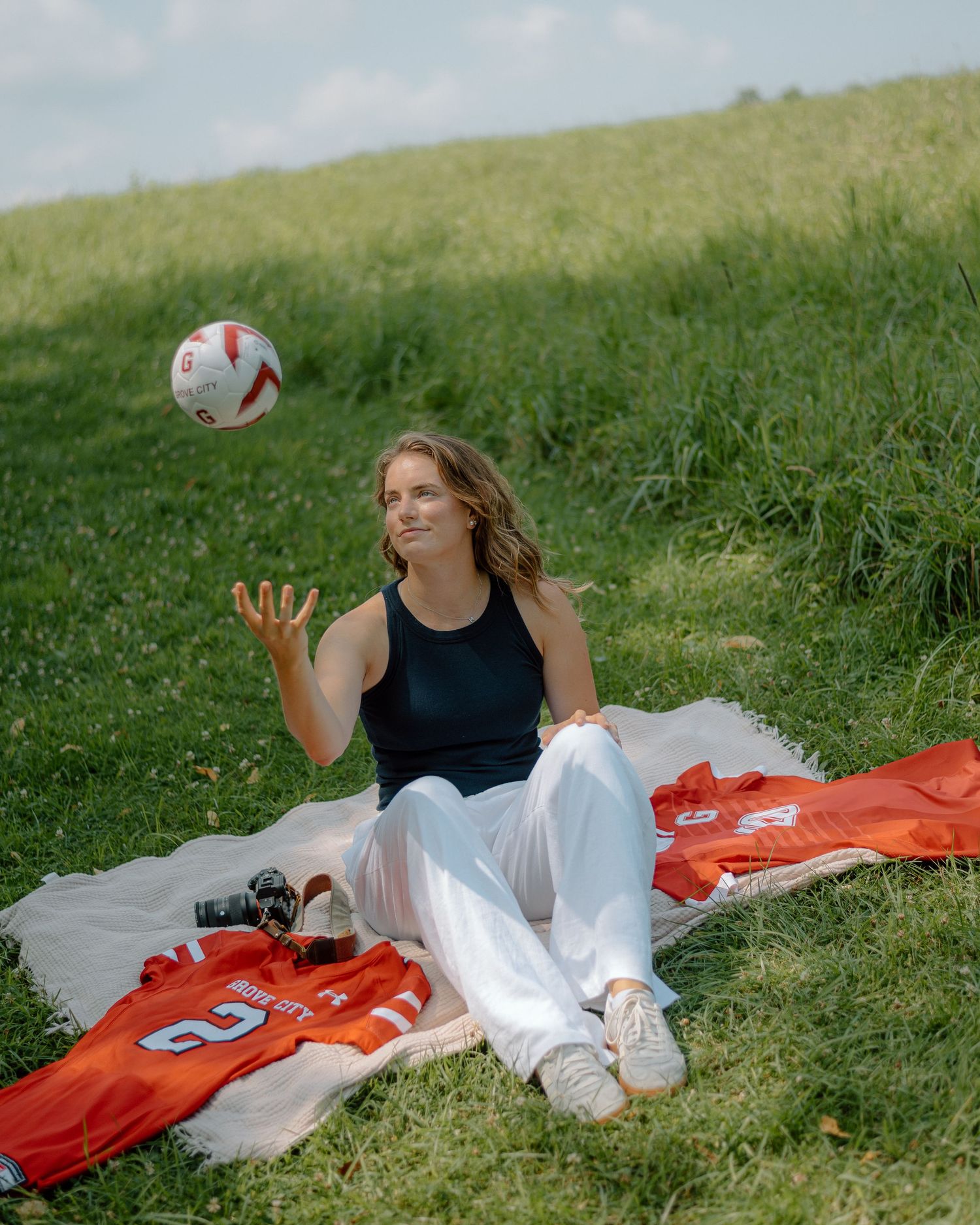 A person in a black top and white pants lies on a red blanket in the grass on a sunny day.