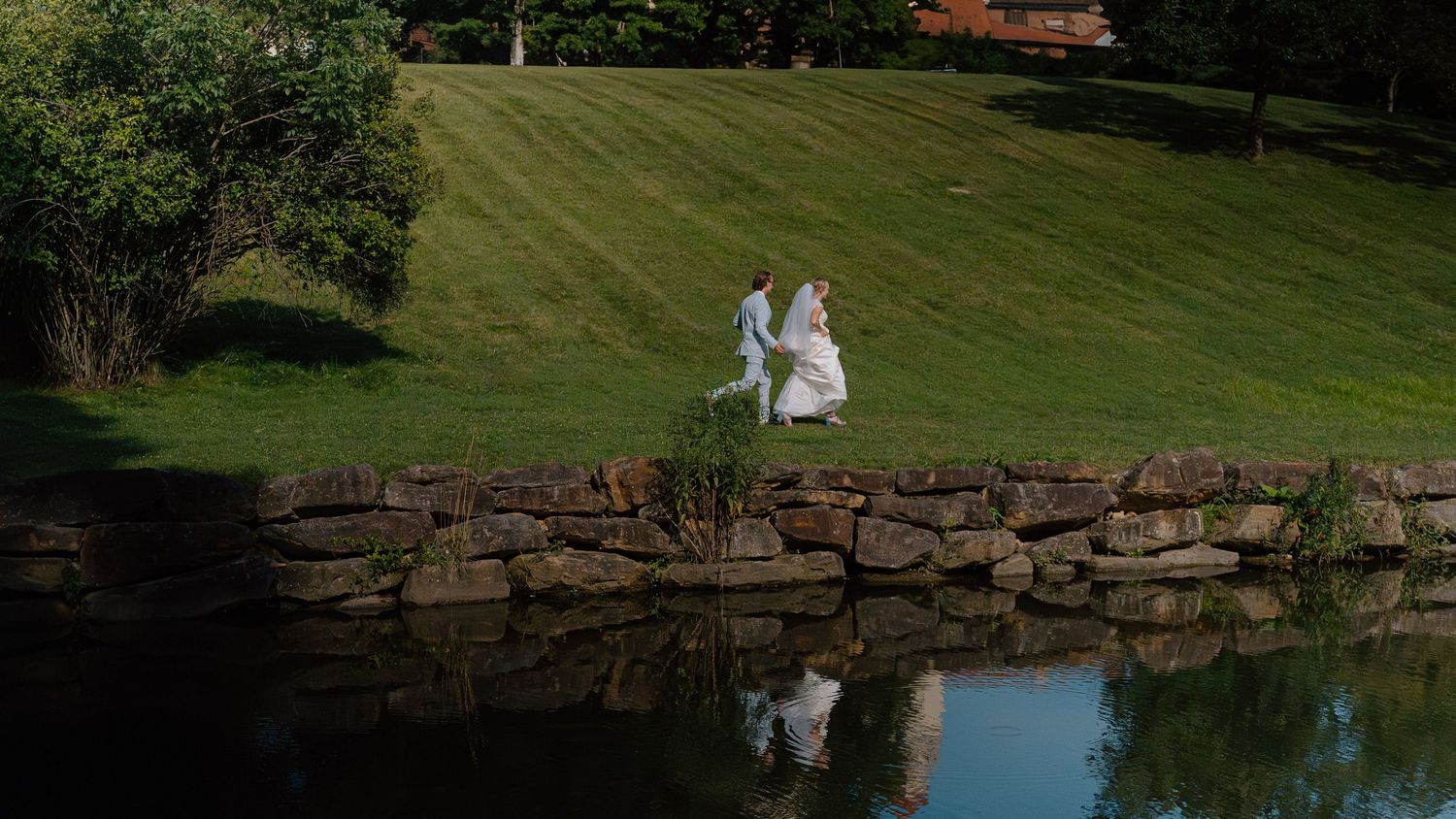 Two people in white gowns walking together on a grassy hillside reflected in a tranquil pond near a stone retaining wall.