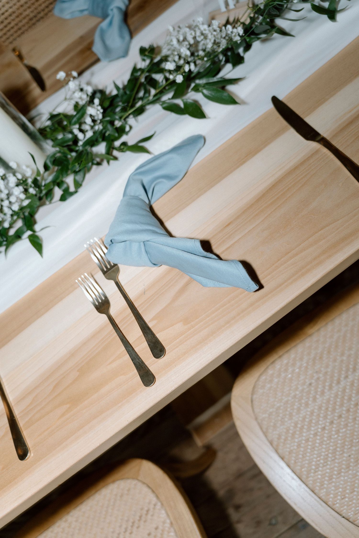 A rustic wooden table setting with light blue napkins, silverware, and delicate greenery garland for an elegant event.