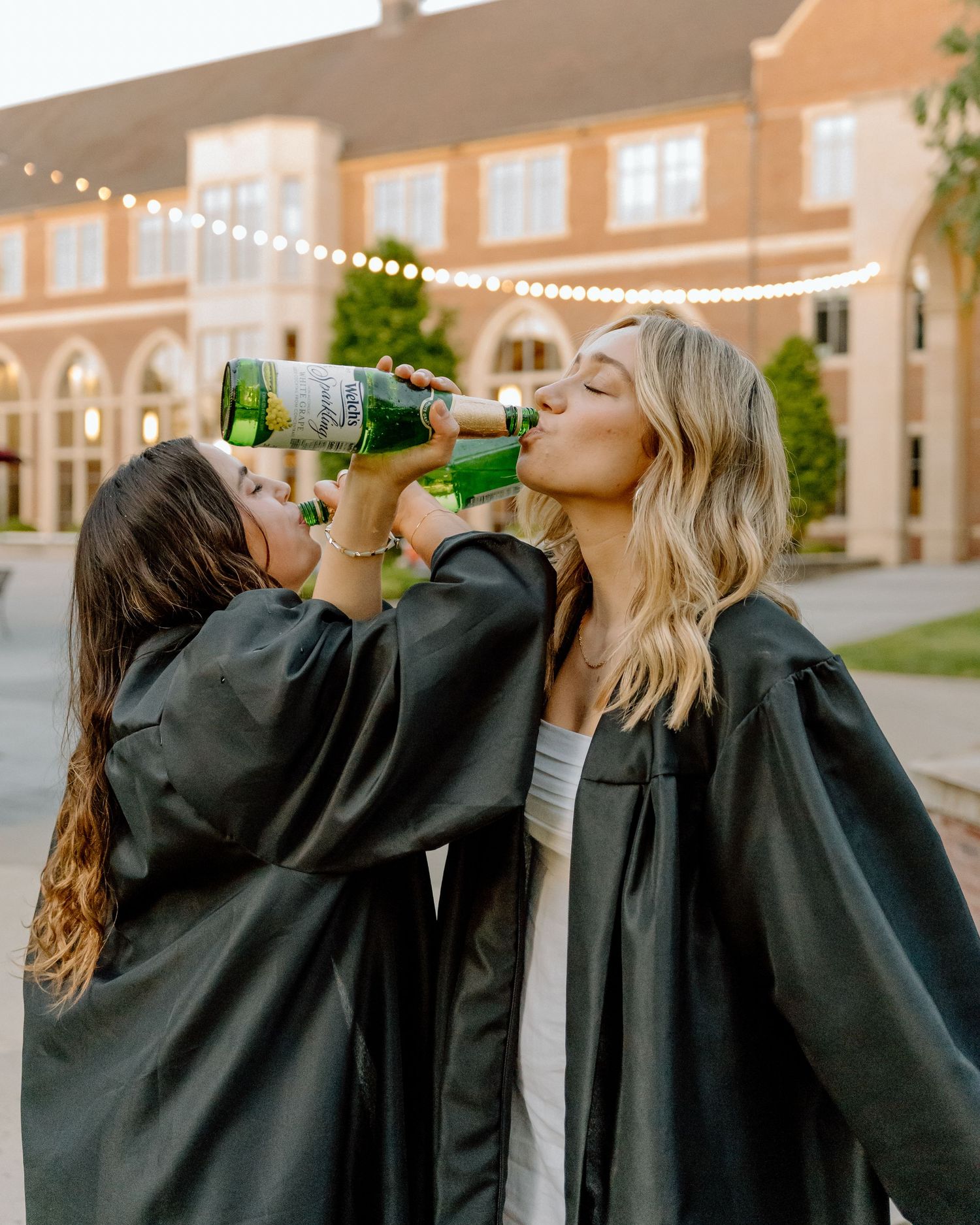 Graduates celebrating in black gowns take celebratory drinks while standing outdoors on campus.