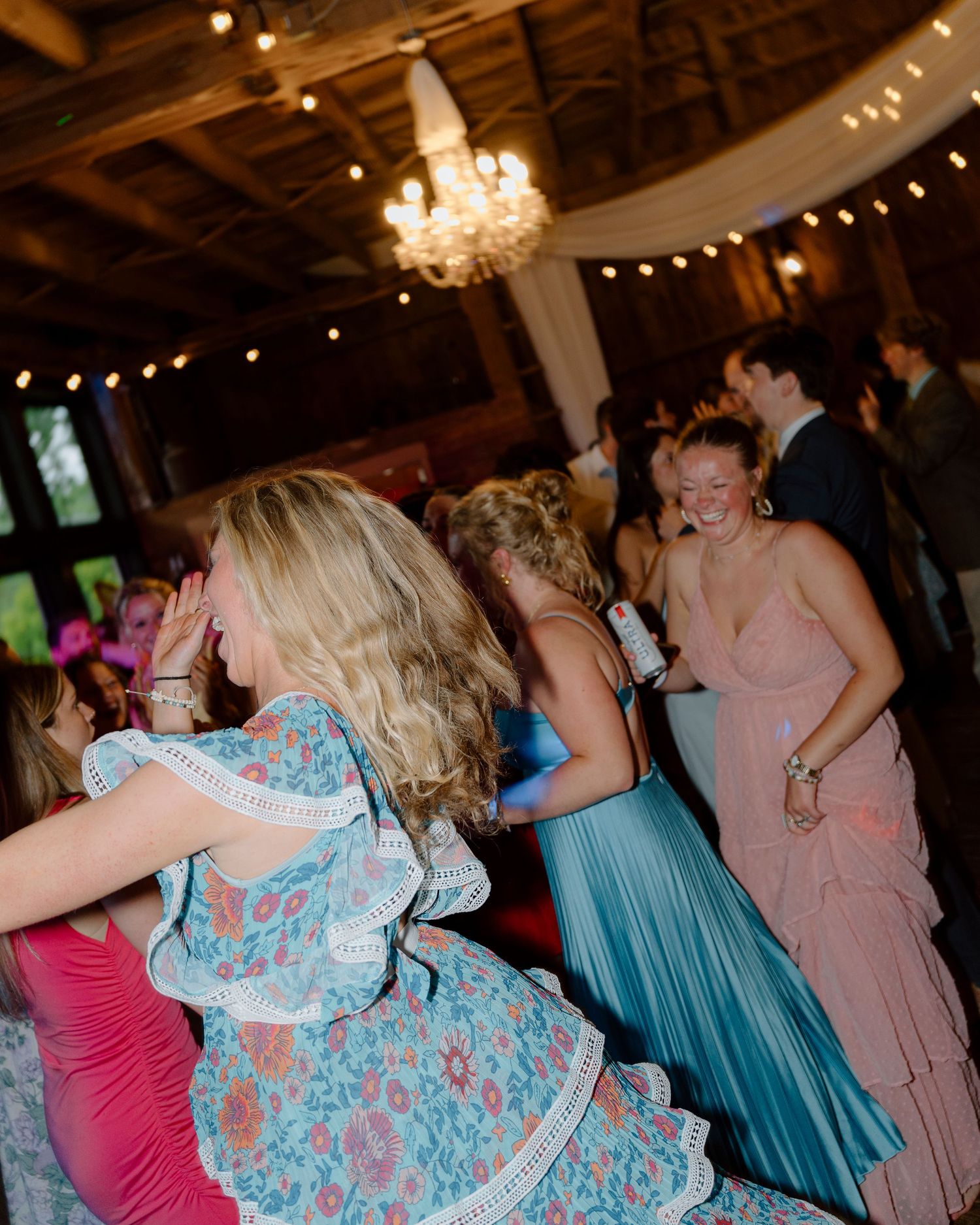 Wedding reception dance floor filled with guests in colorful dresses dancing under chandelier.