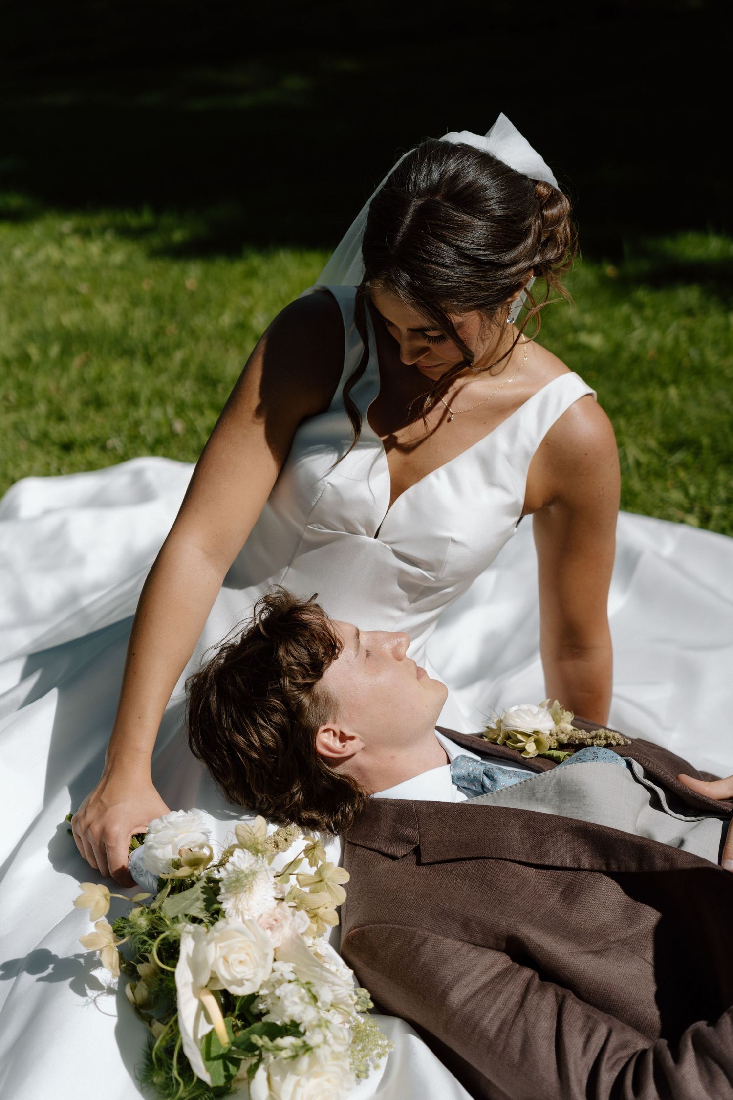Bride in white gown sits beside groom in brown suit lying on grass, sharing a romantic moment outdoors.
