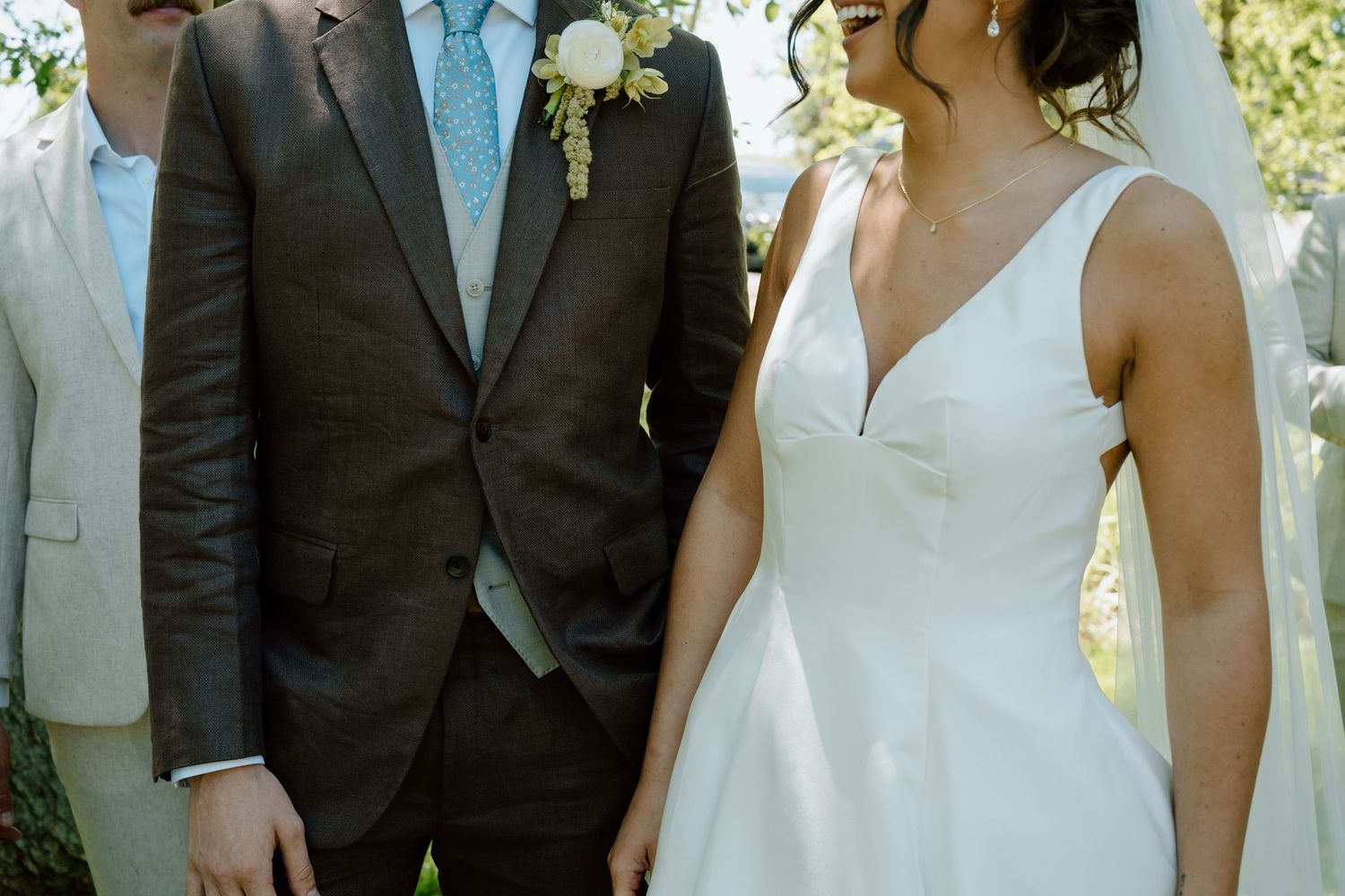 Bride in white V-neck gown stands beside groom in brown suit with floral boutonniere at outdoor wedding.