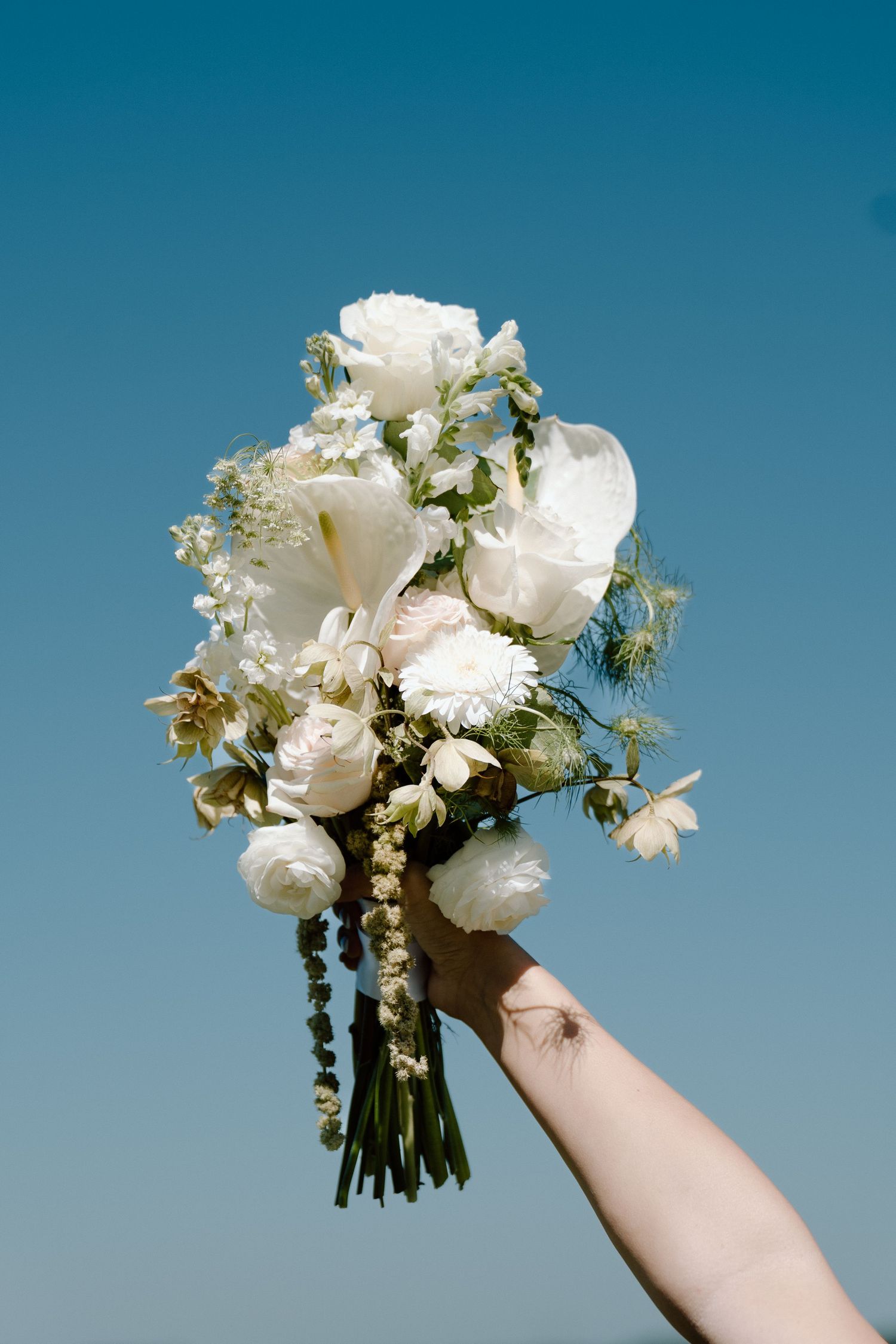 A hand holds a lush white wedding bouquet of roses and mixed blooms against a clear blue sky.