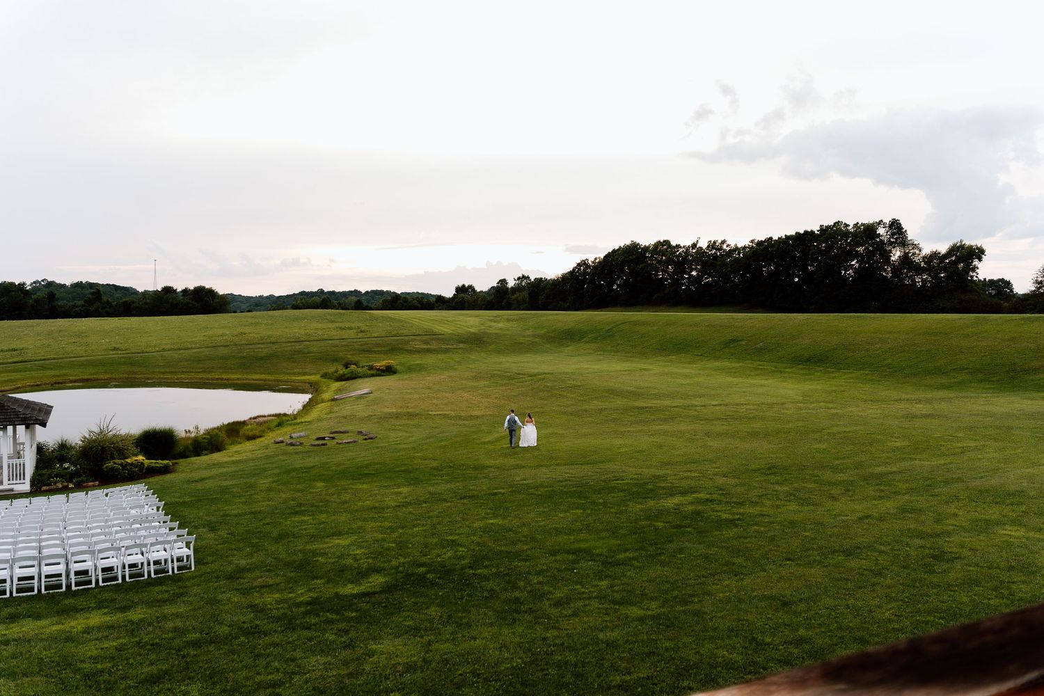 Couple stands in vast green golf course field at dusk, with white chairs arranged for outdoor wedding ceremony nearby.