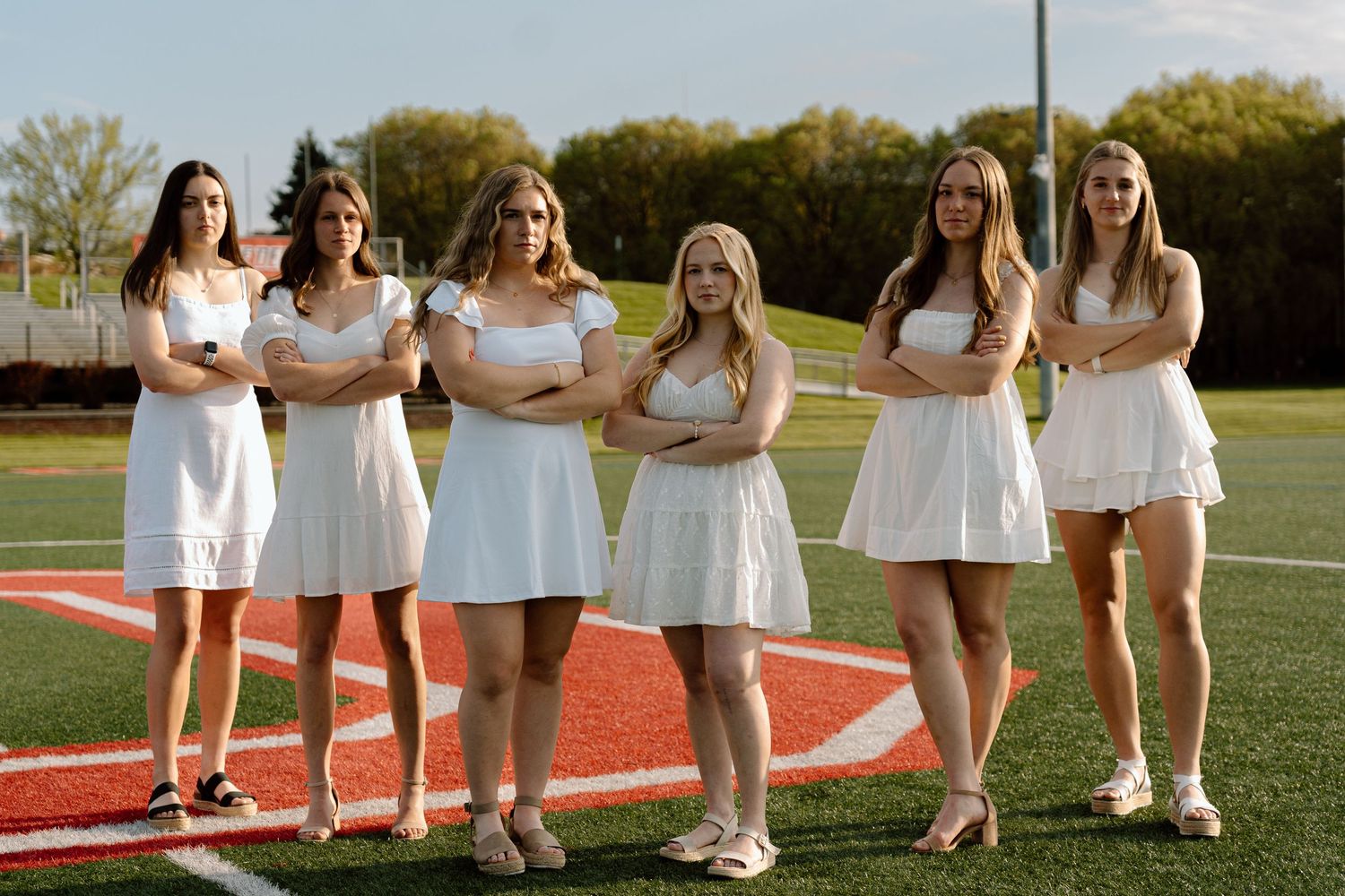 Girls in white outfits pose together on athletic field during sunset photo session.