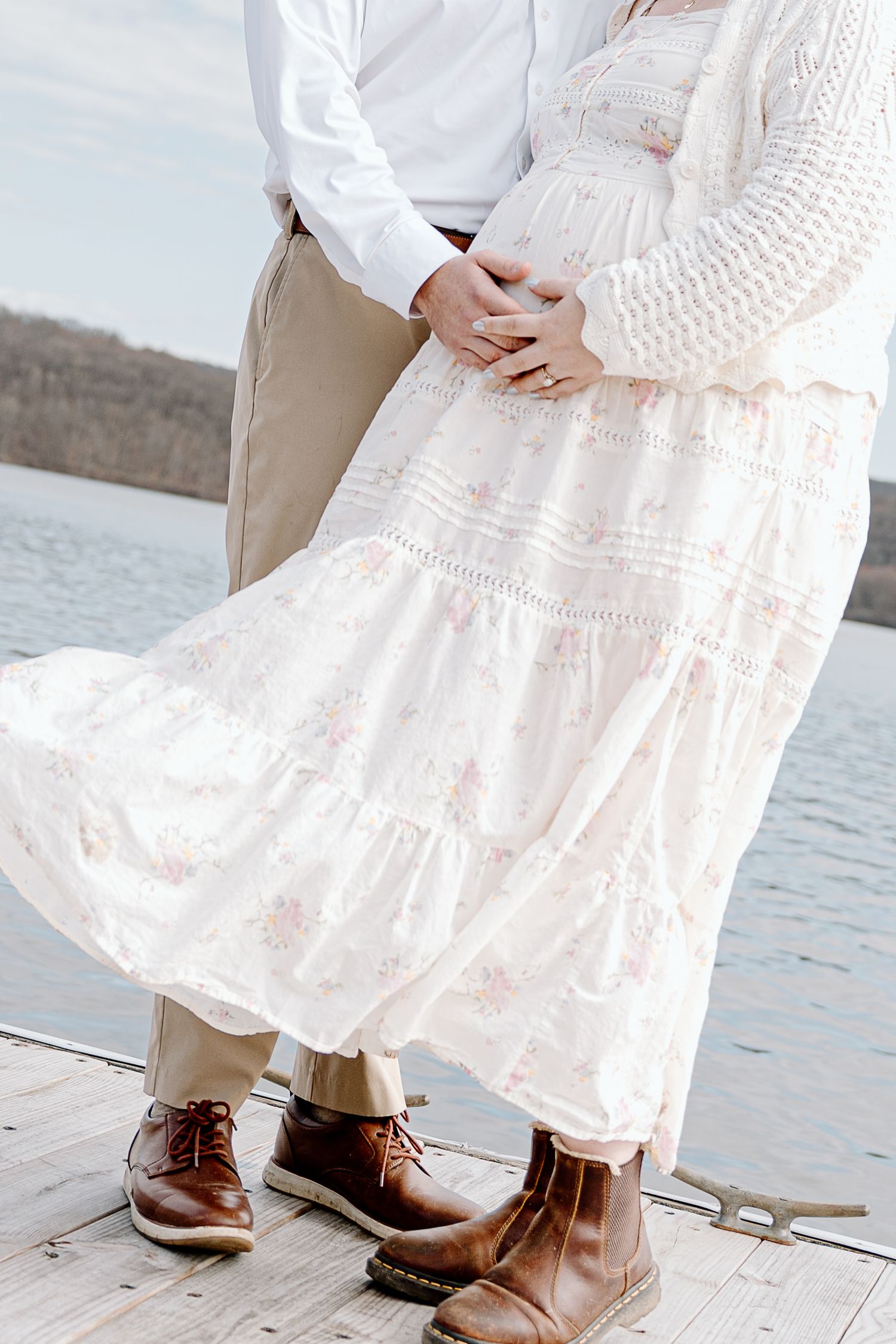 Couple in boho maternity photo on dock, woman in floral tiered dress, man in khakis and white shirt, both wearing brown boots.