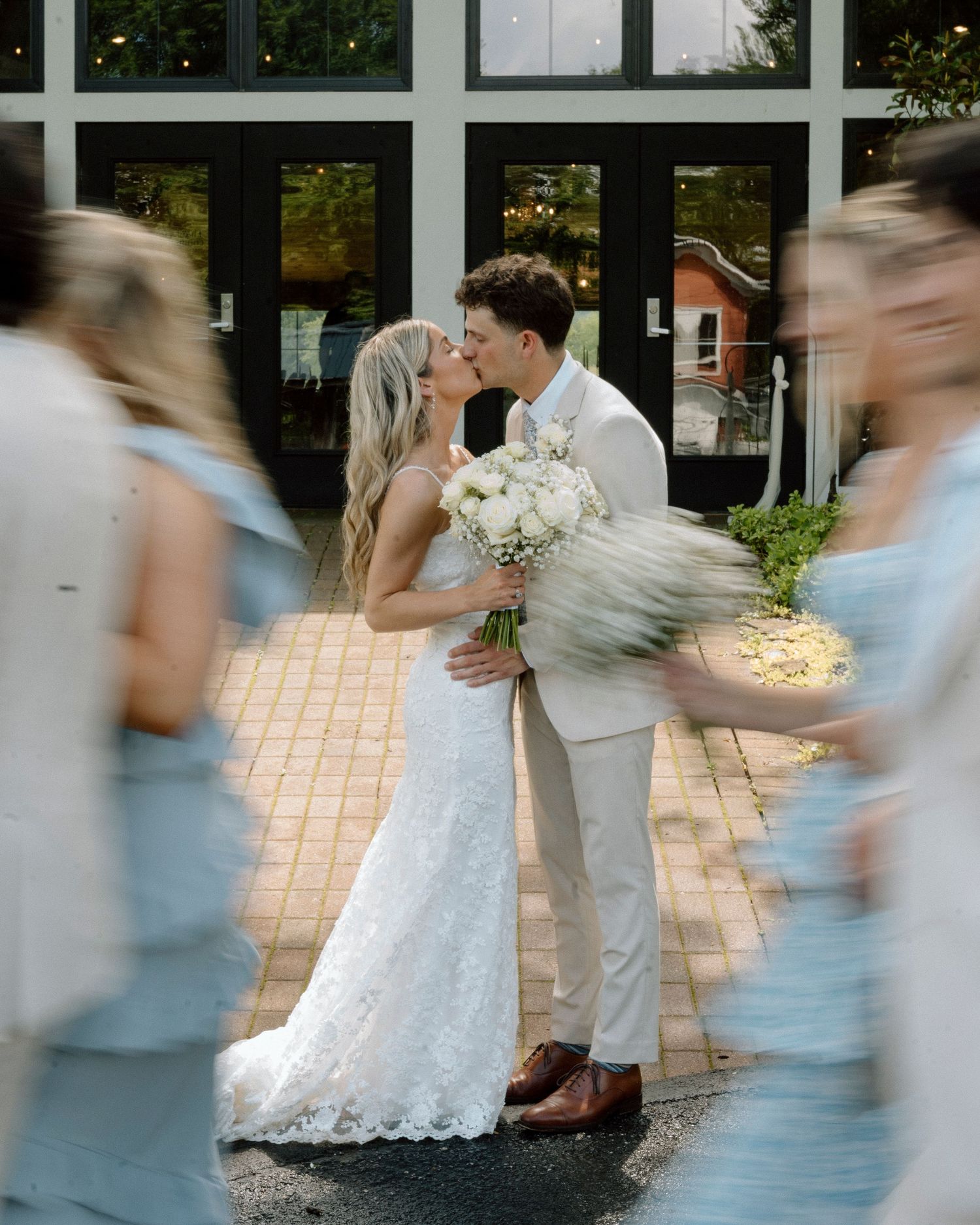 Newlyweds share a romantic kiss on brick pathway while bridesmaids in blue dresses create motion blur around them.