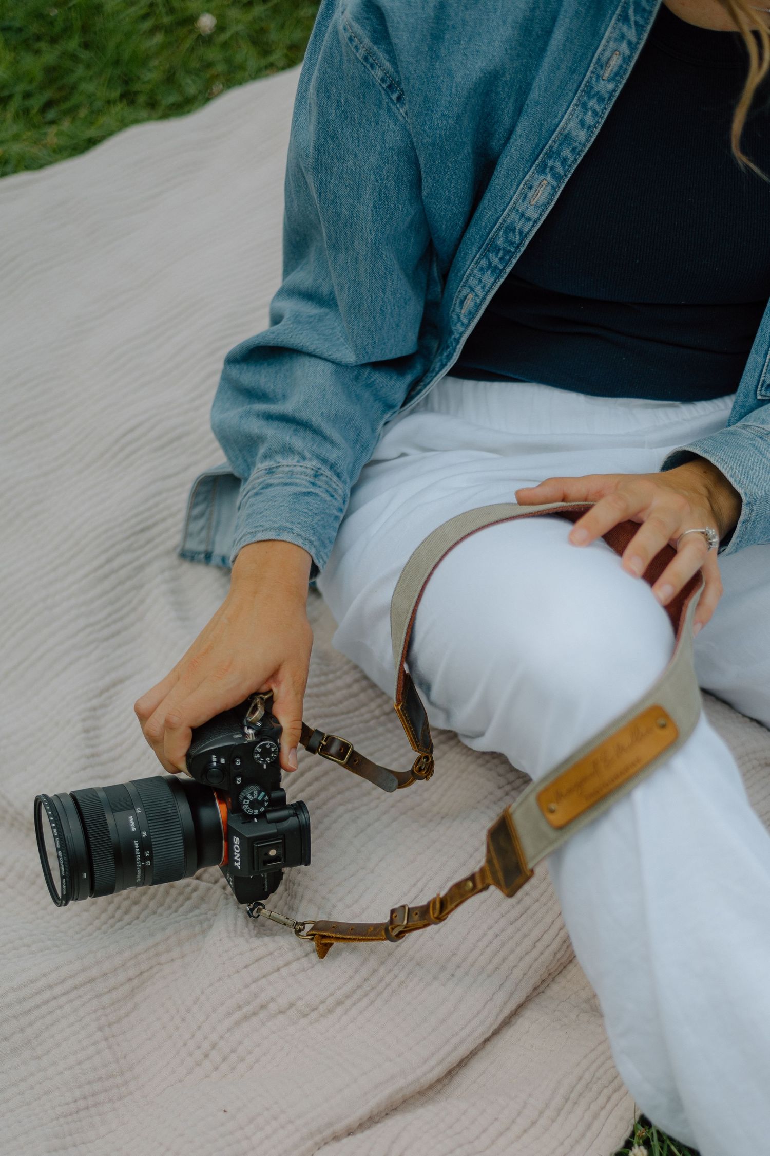 A photographer sits on a blanket with a digital camera and denim jacket, wearing white pants and leather camera strap.