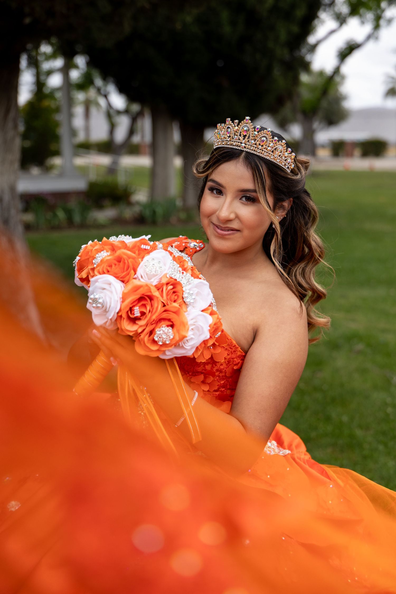 Elegant portrait of woman in flowing orange gown with crown and bouquet in garden setting.