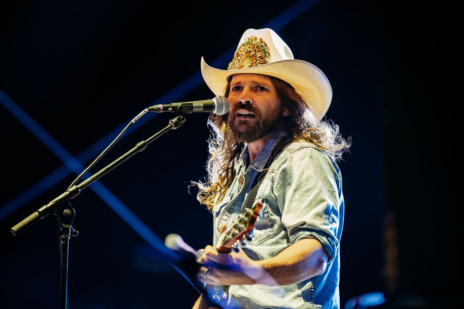 Musician in white cowboy hat performing on stage with guitar and microphone under blue lighting.