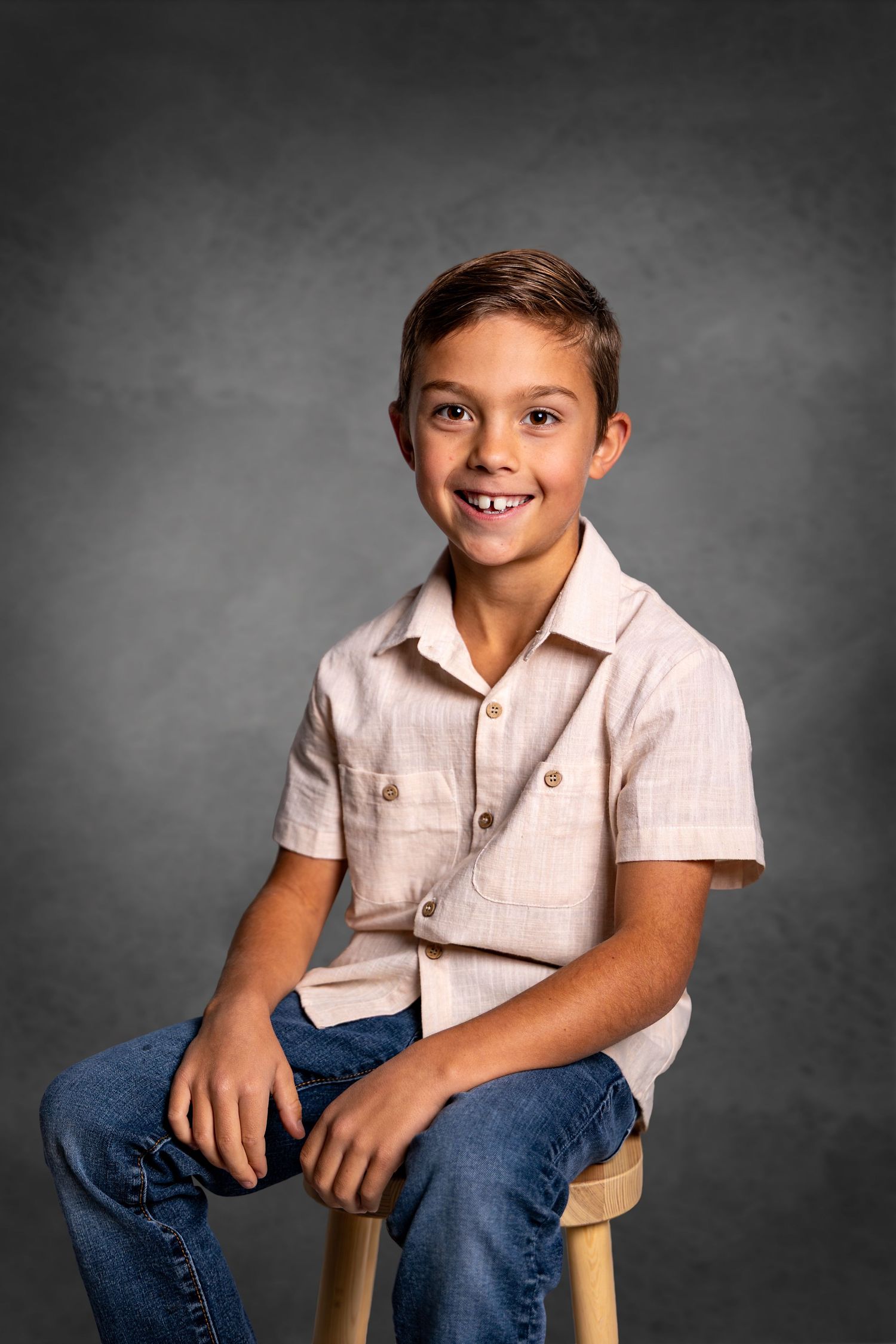 Smiling boy in beige shirt and jeans sits on wooden stool against gray studio backdrop.