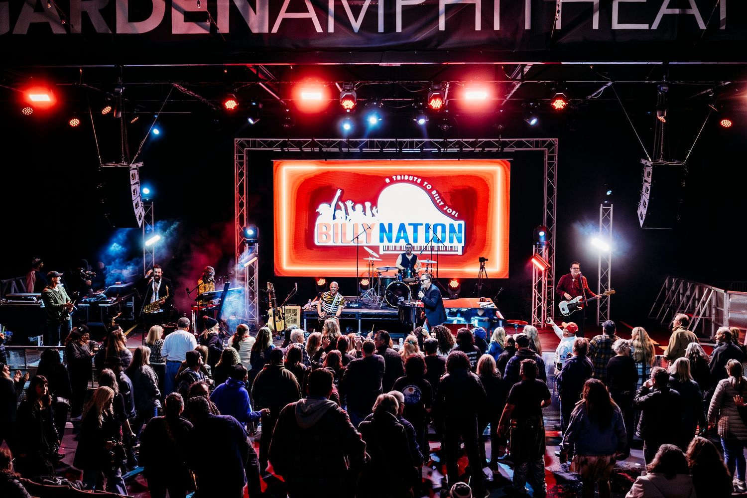 Crowd enjoys outdoor concert at Garden Amphitheater with red lighting and stage signage in Garden Grove.