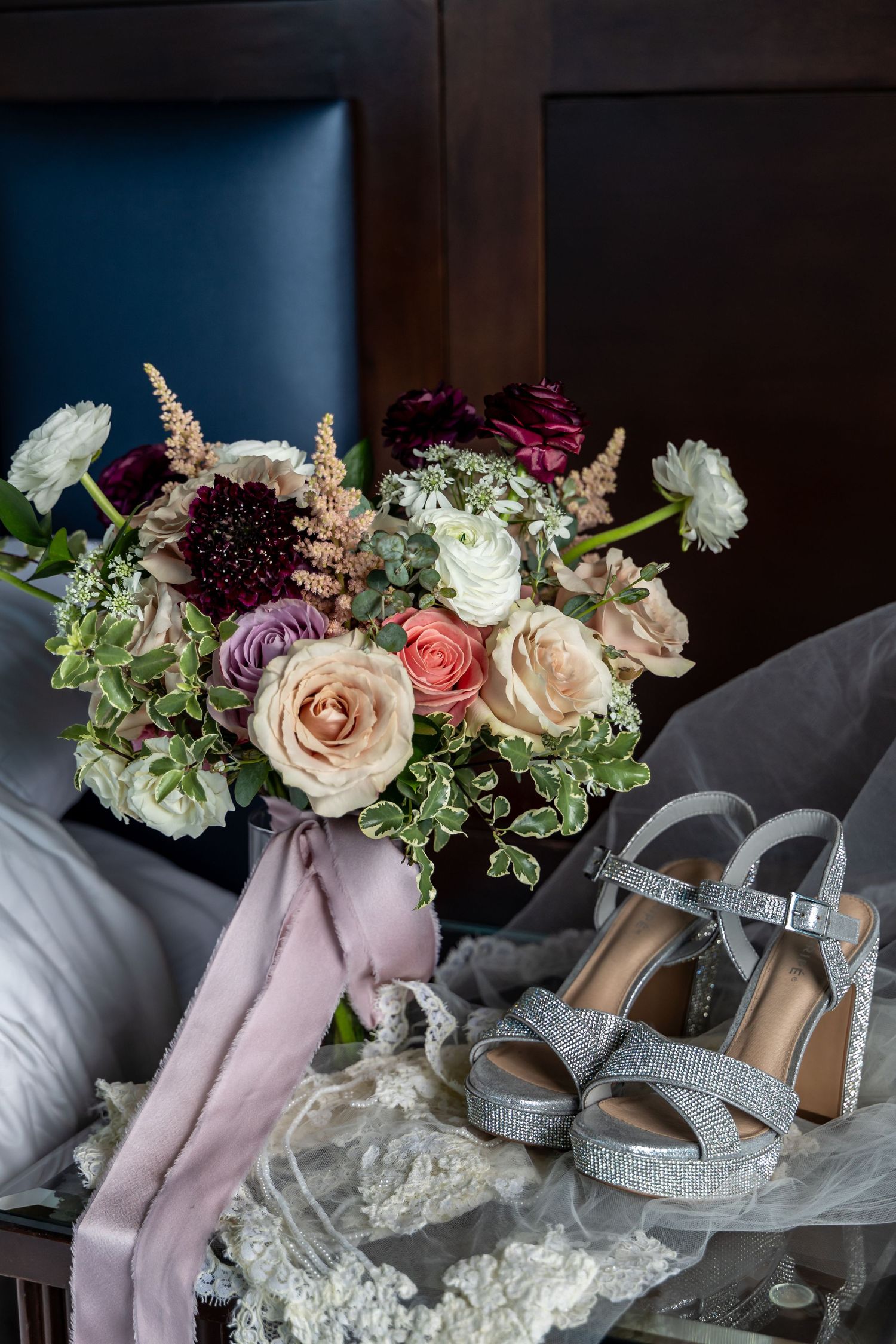 Wedding accessories including silver heels, roses, and silk ribbons arranged on a grey surface.