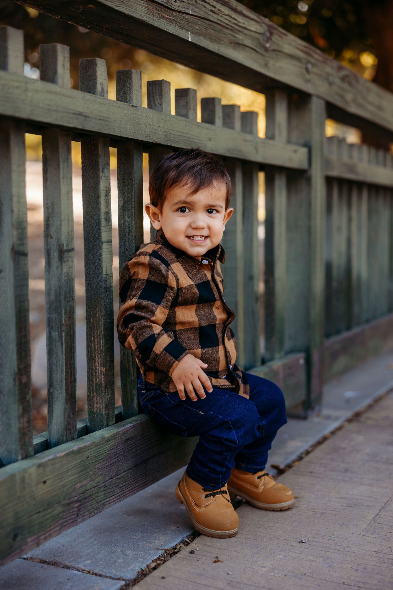Child in plaid shirt and jeans crouches playfully by wooden railing.