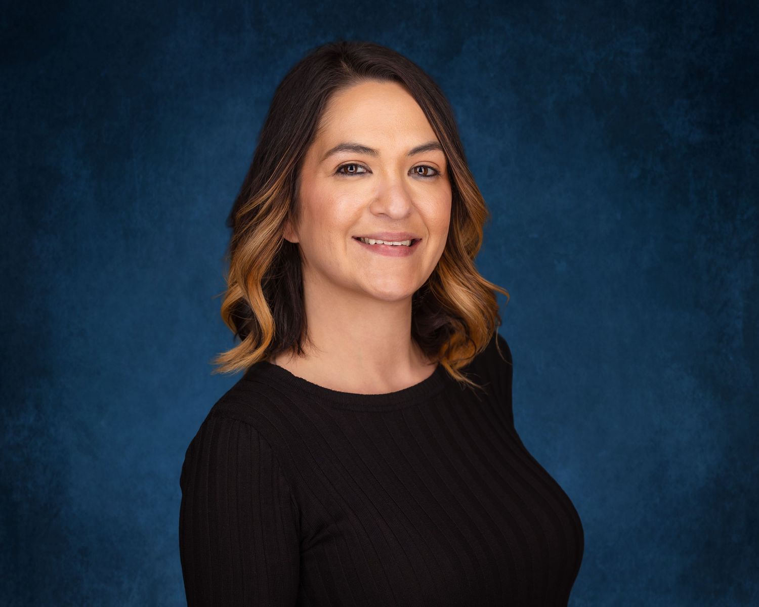 Professional headshot of a smiling woman with brown ombre hair wearing a black top against a blue background.