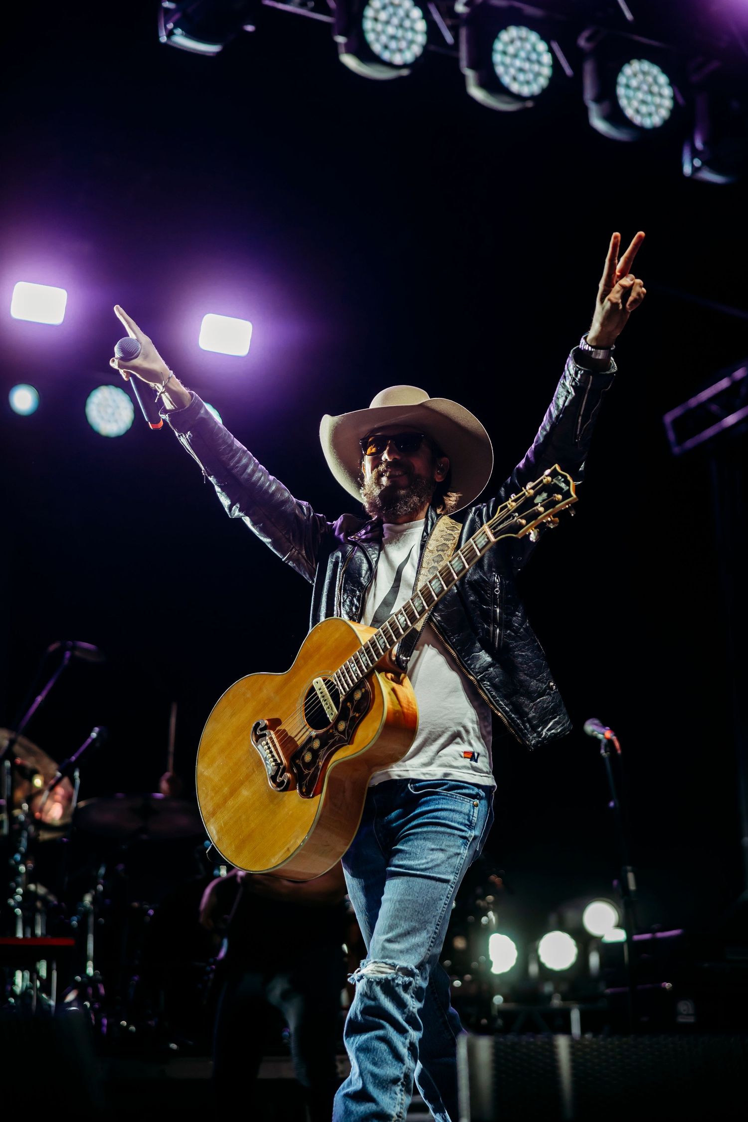 Performer in cowboy hat and denim raising arms in victory pose while holding acoustic guitar on stage.