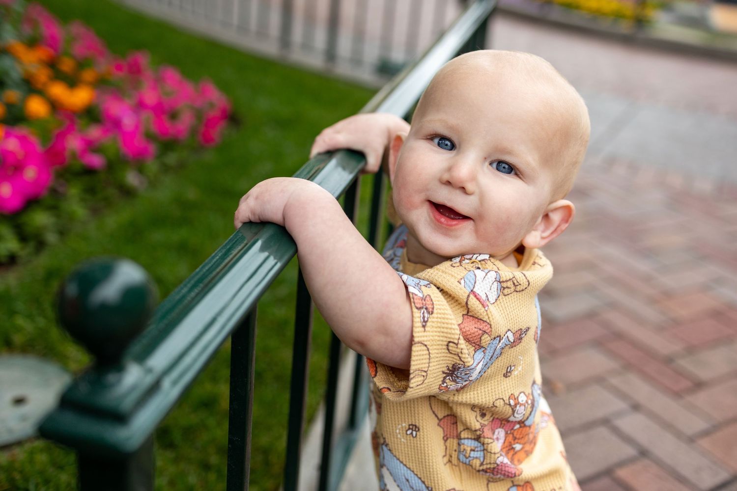 Small child in floral outfit holds onto railing near colorful garden flowers on brick walkway.