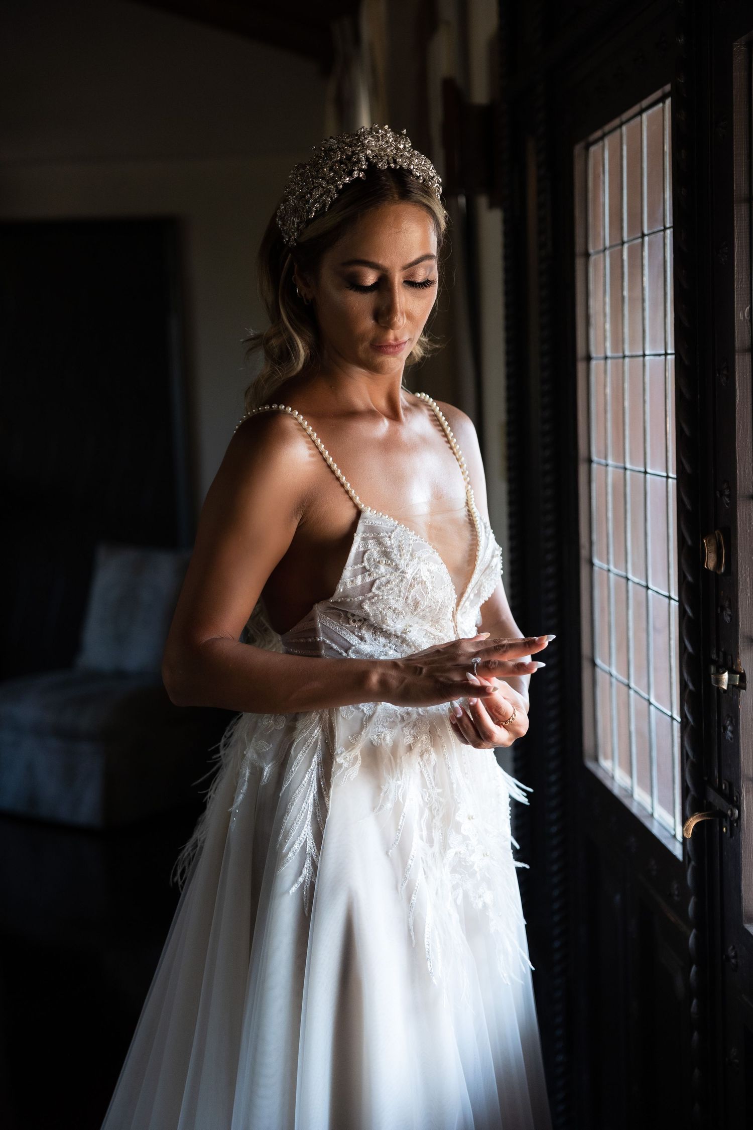 A bride in white lace gown and tiara stands by window in dramatic lighting.