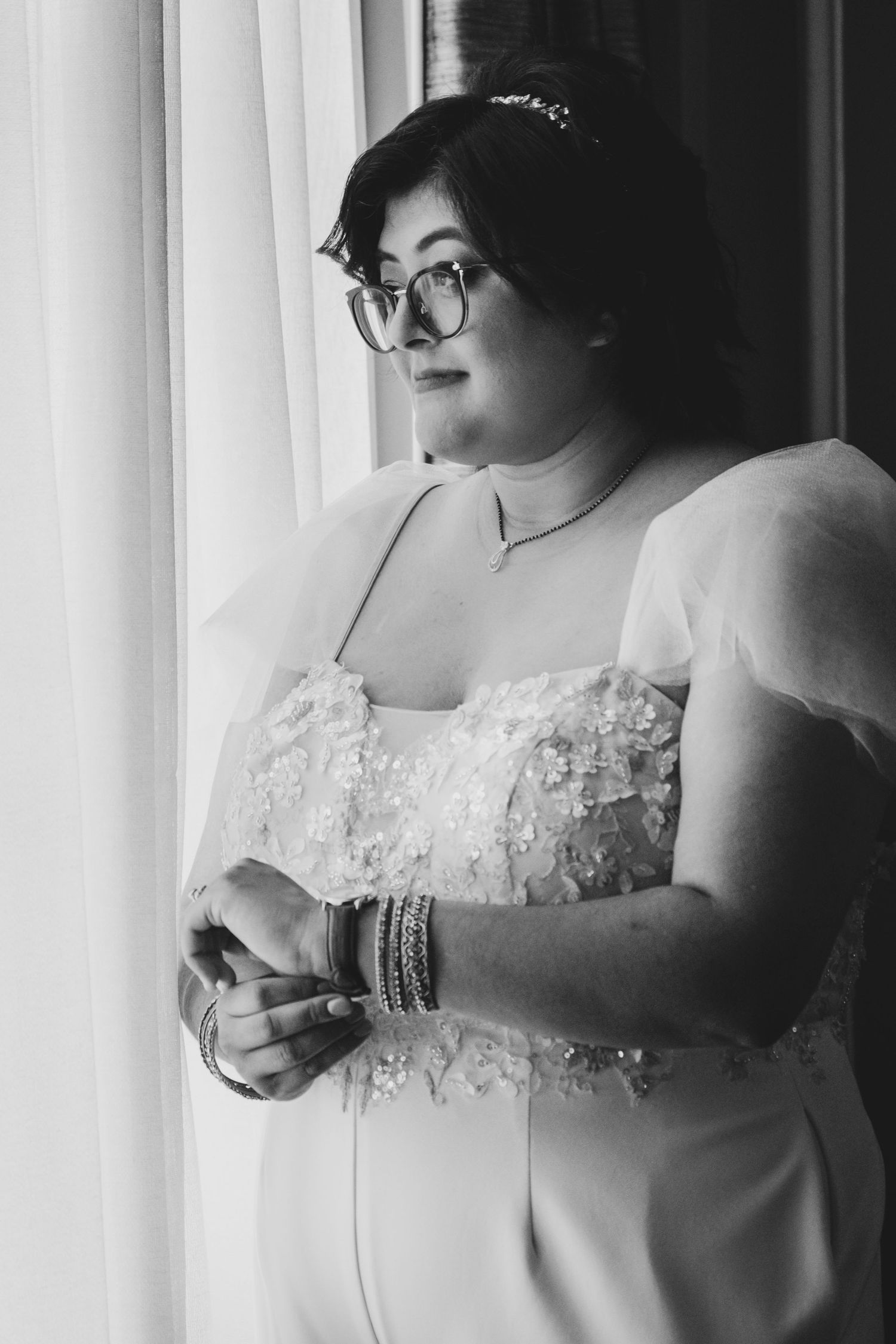 Black and white portrait of someone in a detailed lace wedding dress by a window.