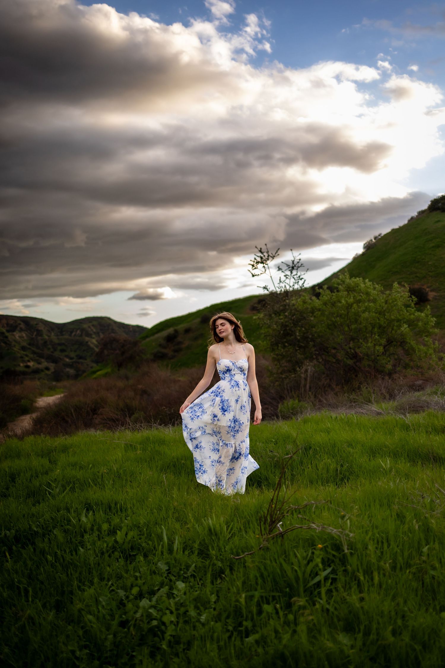 Person in flowing blue floral dress standing on hillside under dramatic cloudy sky in Redlands.