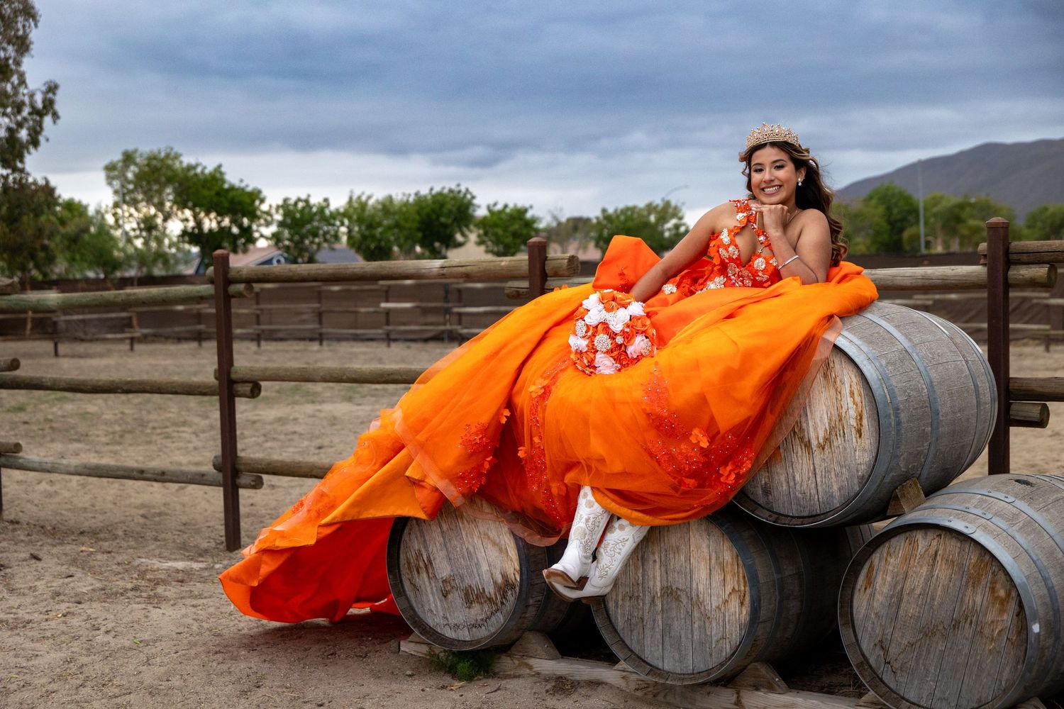 A bright orange formal dress contrasts against wine barrels in a rustic outdoor setting.