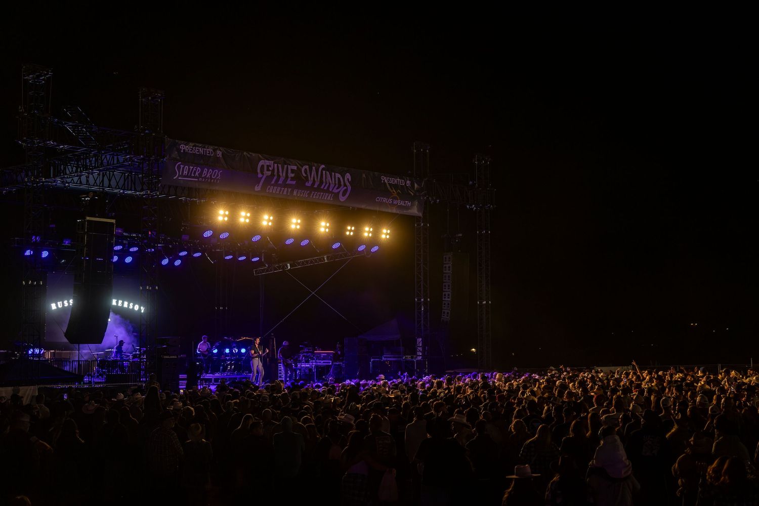 Concert stage lights illuminate a large crowd at an outdoor music festival.