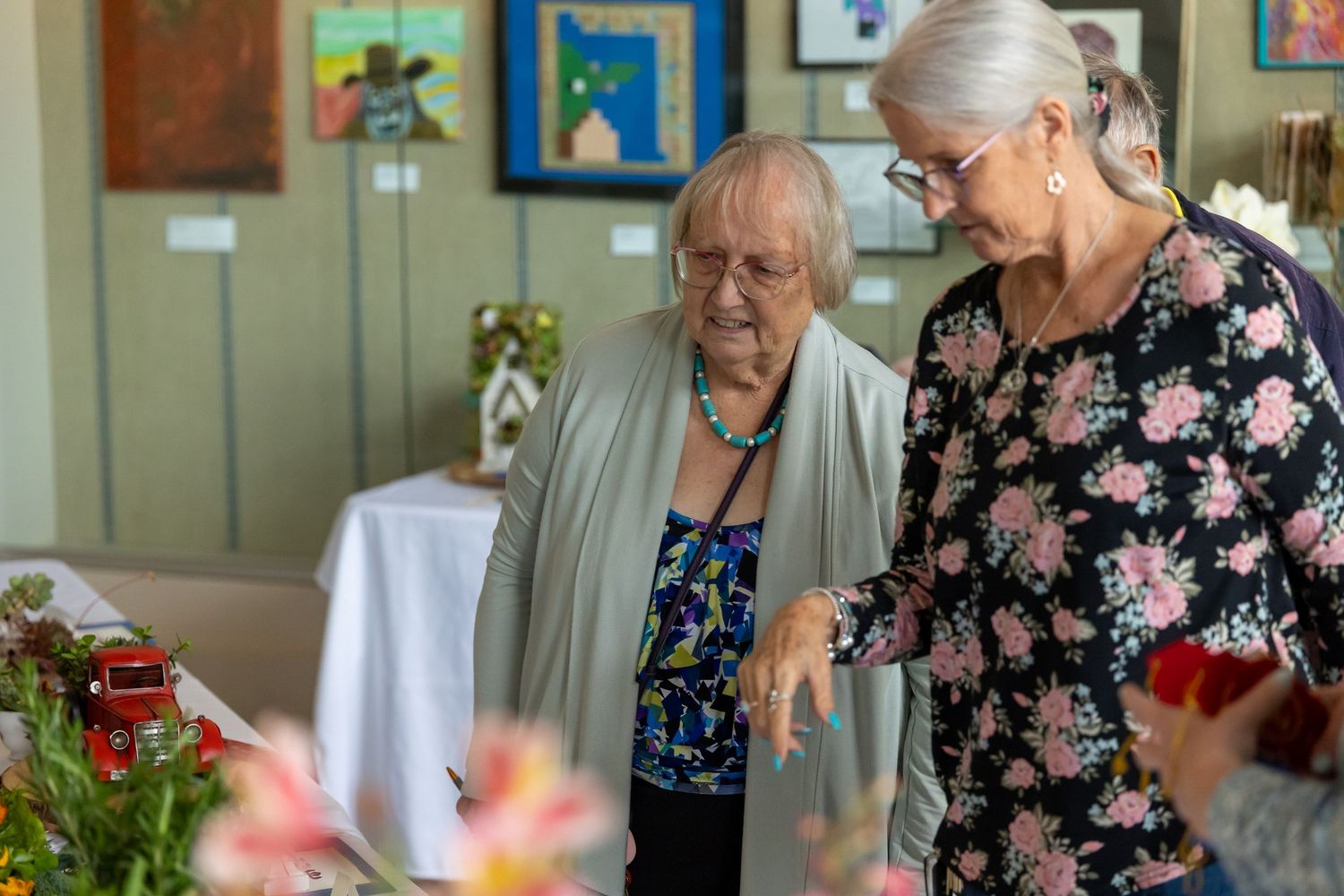 Two elderly women looking at floral displays in an art gallery.