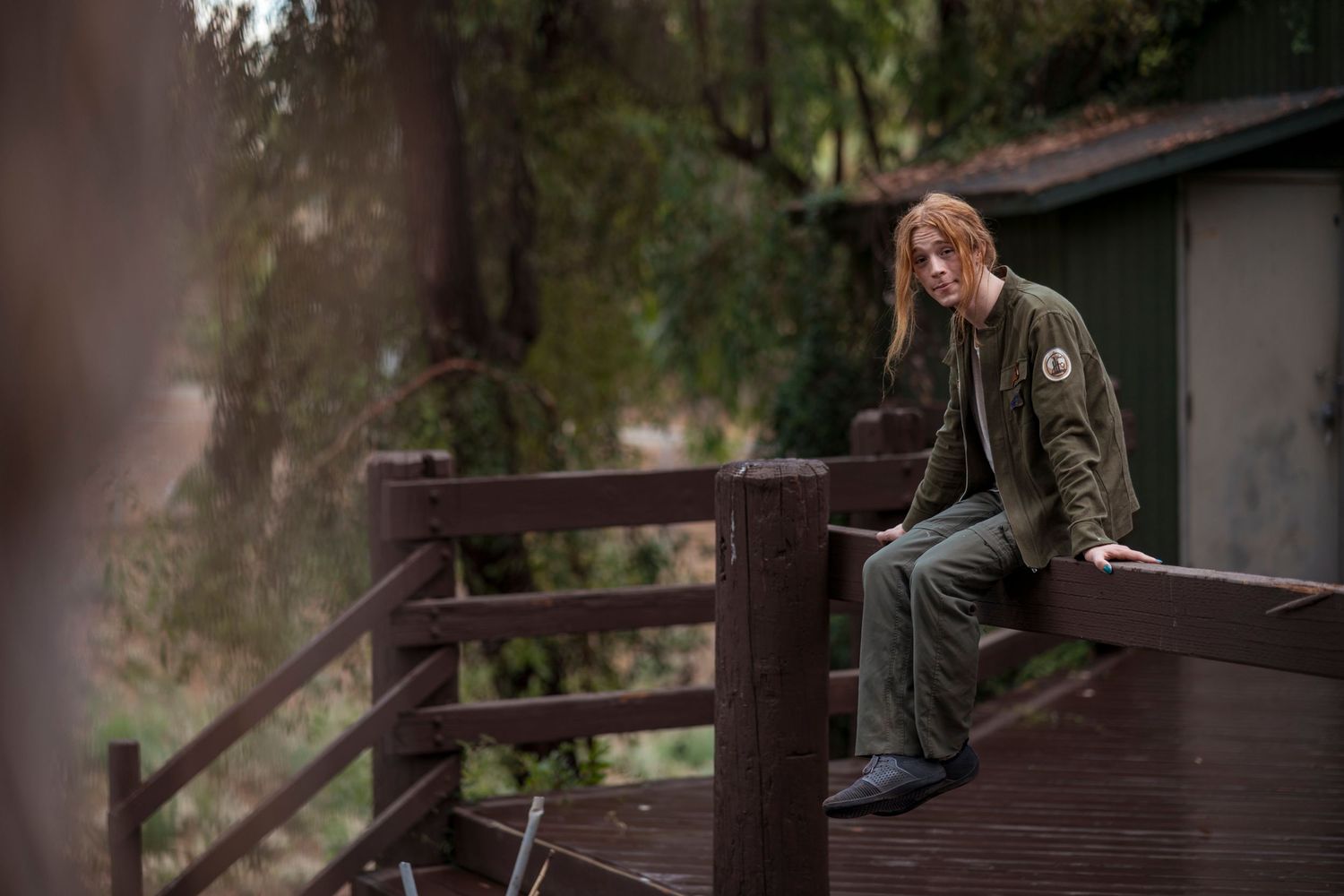 A Graduate sits casually on a wooden deck railing in an olive-green jacket against a forested backdrop in Redlands.