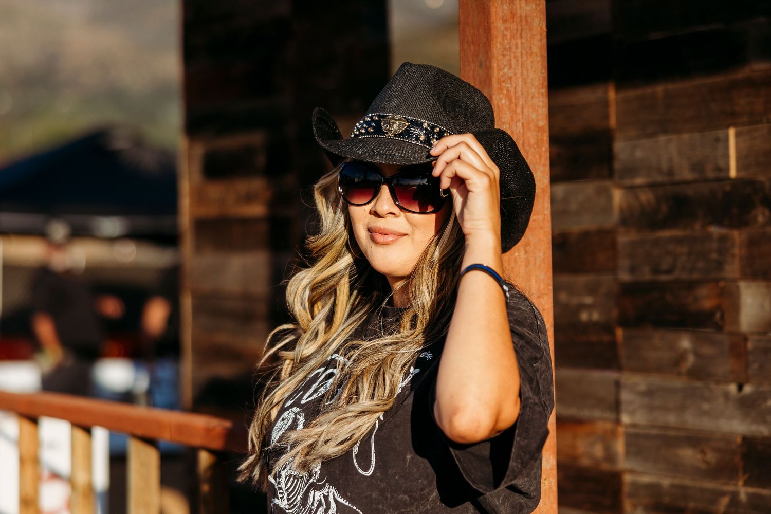 Person in black tank top and sunglasses poses outside rustic wooden building in sunlight.