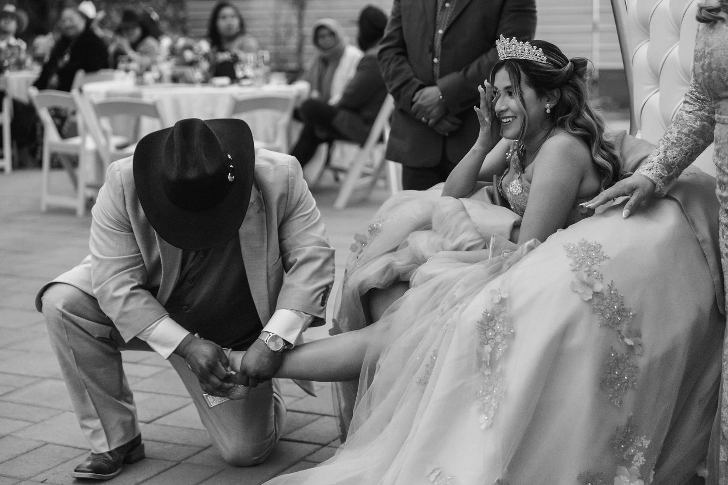 Kneeling figure in formal attire placing a shoe on someone wearing an ornate white dress during a special celebration.