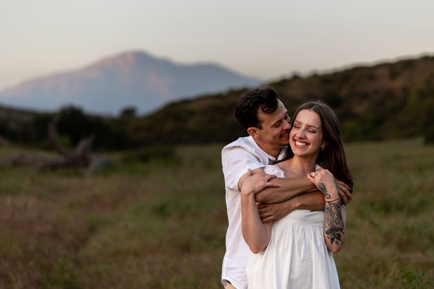 A tender embrace shared between a couple in white clothing against a mountain backdrop.