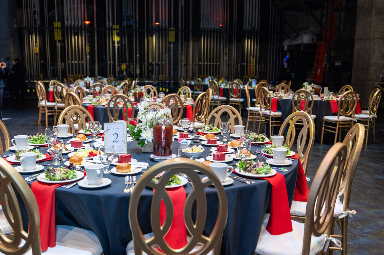 Elegant banquet tables set with red and navy linens, gold chairs, and formal place settings in a dimly lit venue.