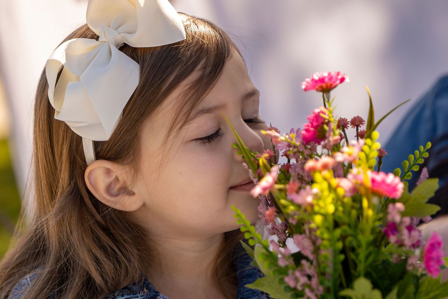 A young girl with a white bow in her hair smells a bouquet of pink and yellow flowers with eyes closed.