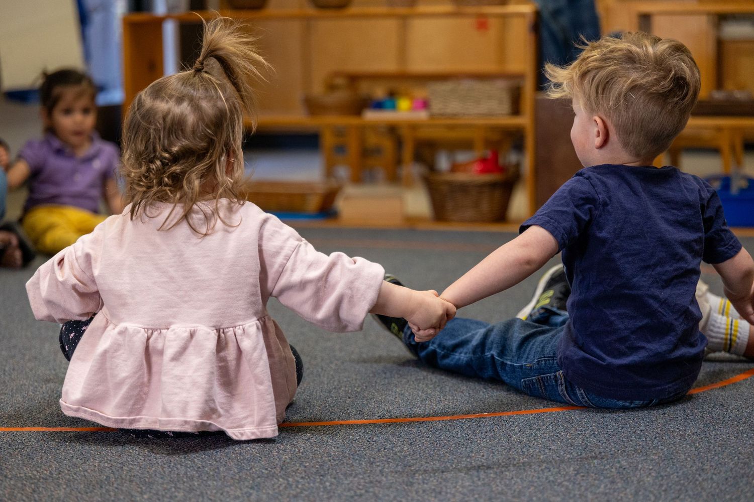 Two young children sitting together on a carpeted floor holding hands in a classroom setting.