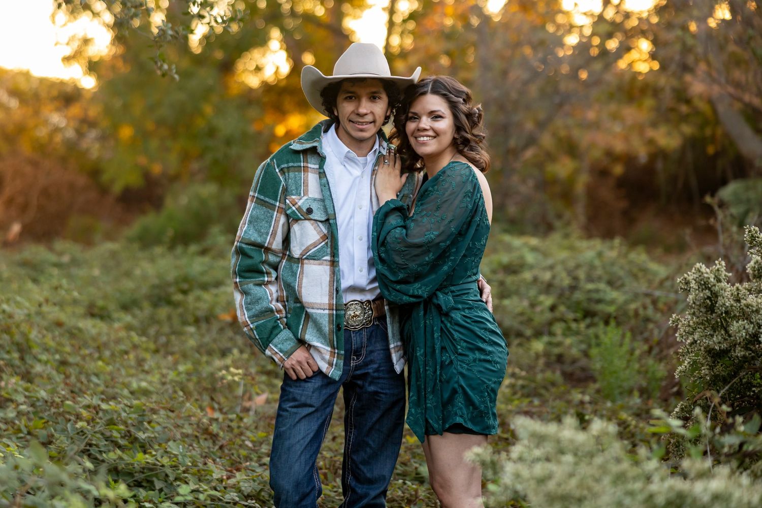 A couple poses together in an autumn setting with warm golden sunlight filtering through trees in the background.