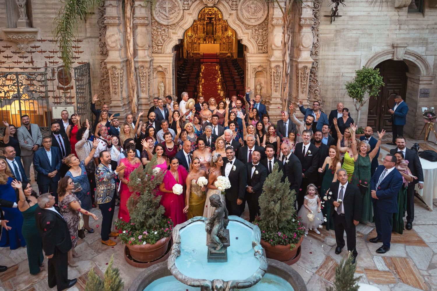 Large wedding group photo outside historic church building with fountain.