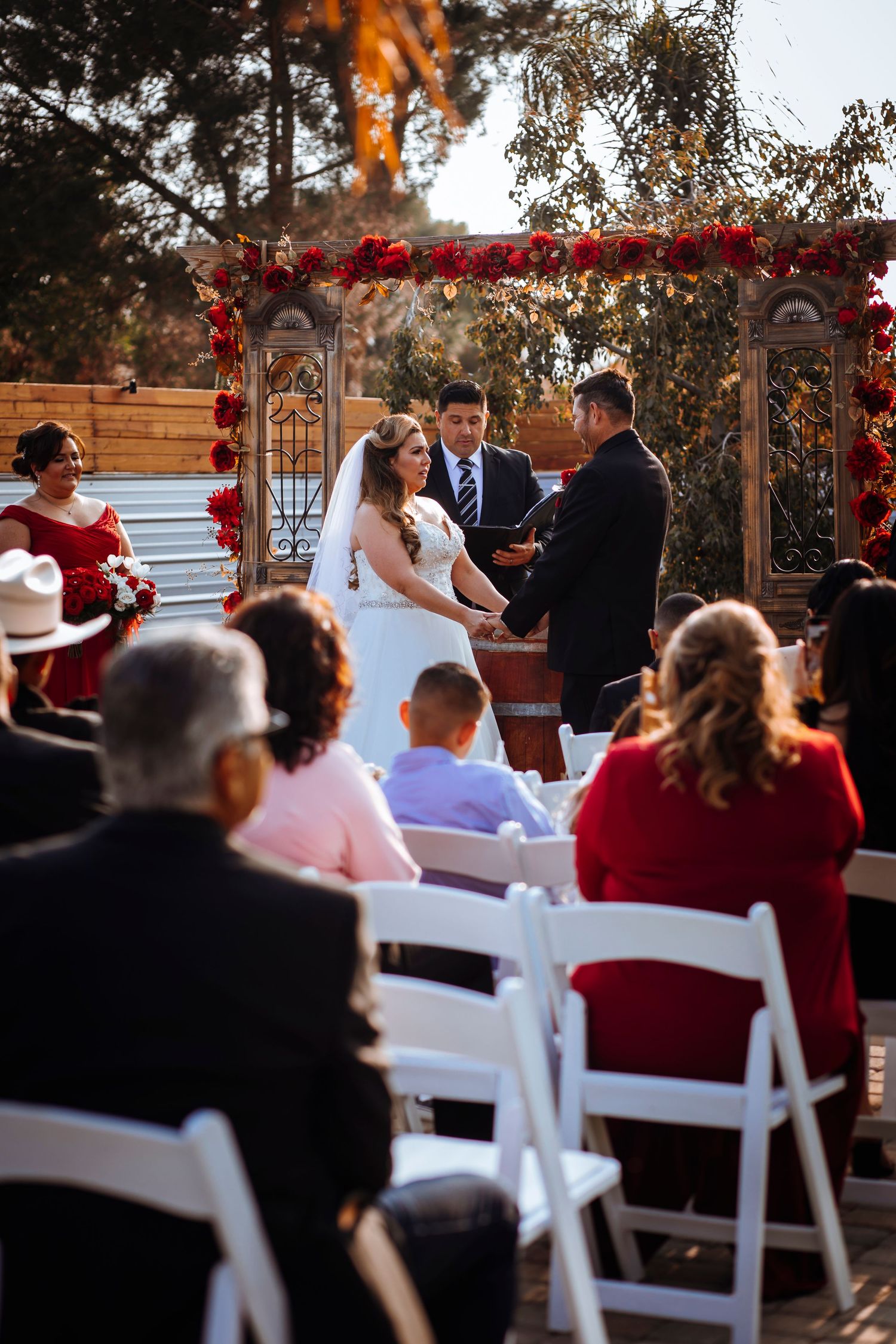 Wedding ceremony setup with white chairs and wooden arch decorated with red flowers at an outdoor venue.